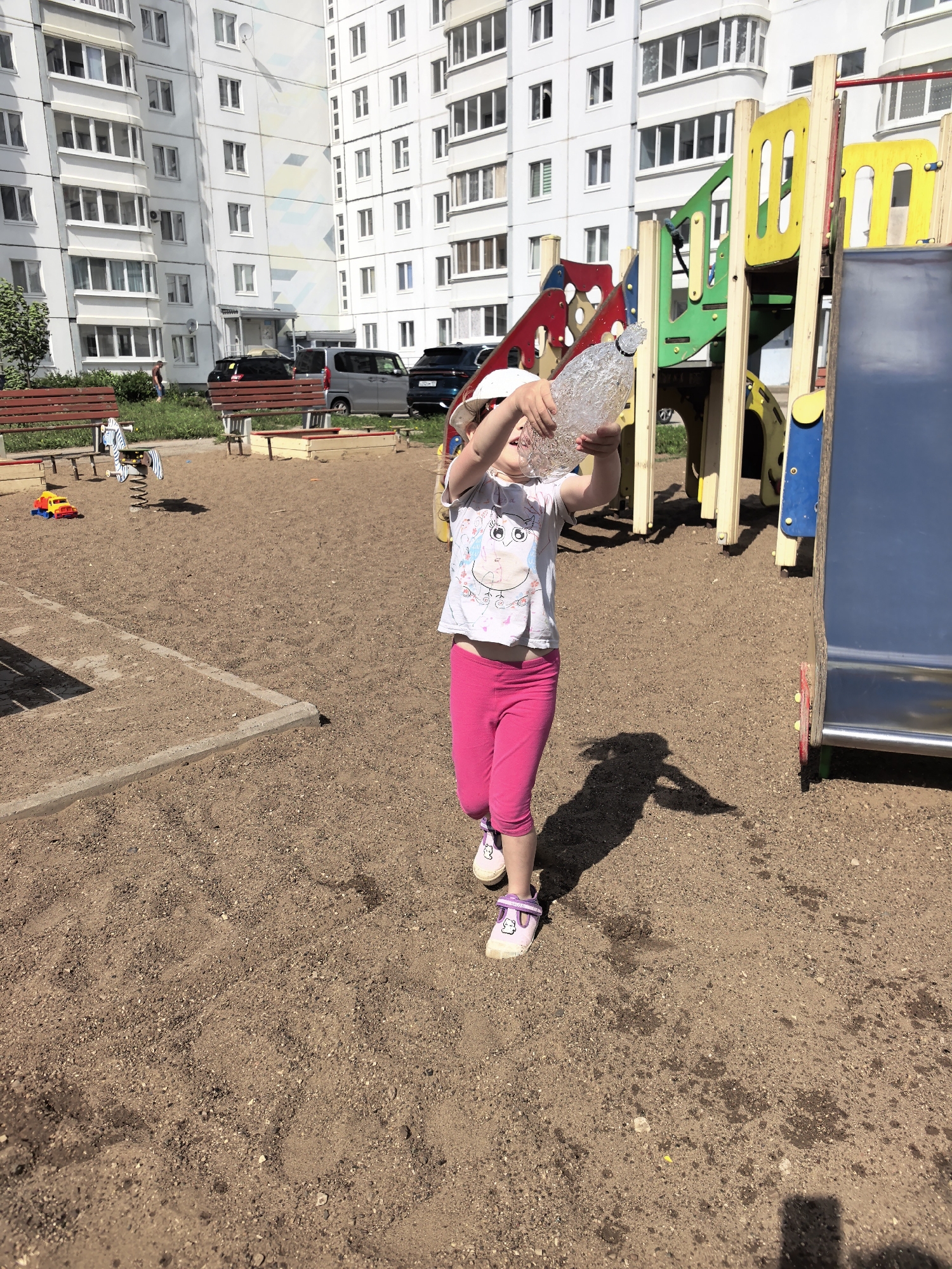 The image shows a young girl in a playground. The playground is located in Фролы, Russia, as indicated by contextual clues. The girl is holding a large, clear plastic bottle filled with water above her head. She is wearing a white t-shirt with a cartoon design, bright pink leggings, and light purple shoes with cartoon characters on them. She also has a white cap on her head. Her shadow is visible on the ground next to her.
The ground is covered in sand. Behind her is a colorful playground structure with a slide, ladders, and other play elements. In the background, there is a multi-story white apartment building with many windows. There are several cars parked near the building, and some trees and benches are visible in the grassy area adjacent to the playground. A small toy truck and a spring rider are also visible in the background. The weather appears to be sunny, as indicated by the distinct shadows.