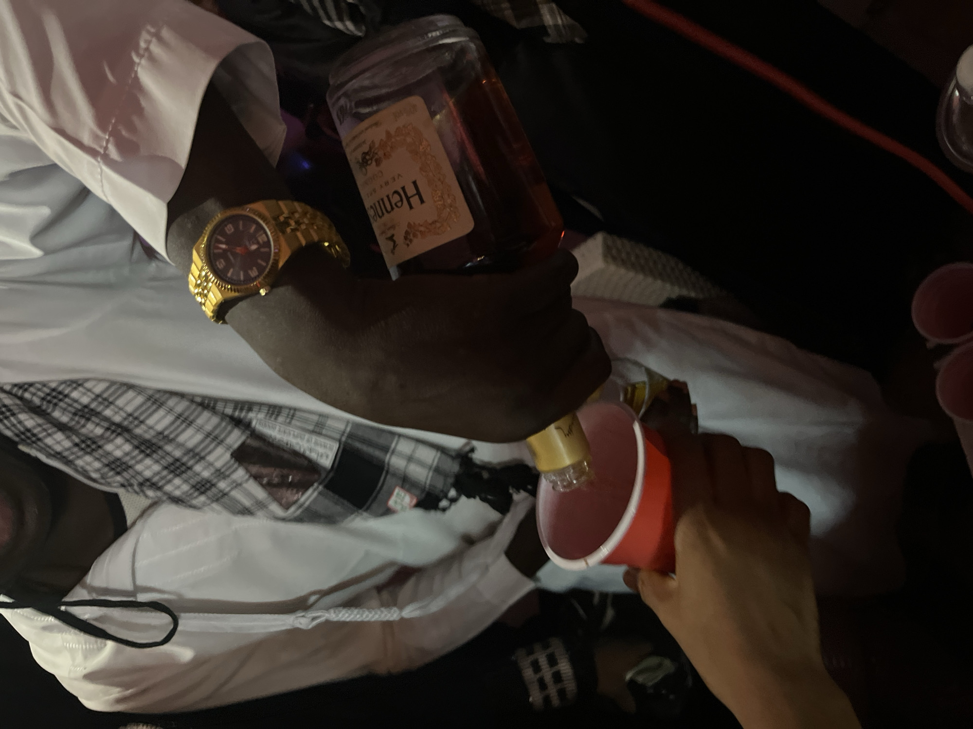 The image displays a close-up, angled view of an individual pouring an alcoholic beverage in an indoor setting within Unknown, Unknown. A dark-skinned arm, wearing a gold-colored wristwatch with a dark face and red accents, holds a clear glass bottle of "Hennessy V.S. Very Special Cognac." The amber liquid from the bottle is being poured into a red plastic cup, held by a lighter-skinned hand. The person pouring is wearing a white long-sleeved garment. In the upper left background, a portion of another individual is visible, wearing a white garment with a black cord and a black and white plaid scarf. The scene is dimly lit, with dark background elements and hints of other red plastic cups and a red cable in the lower foreground.