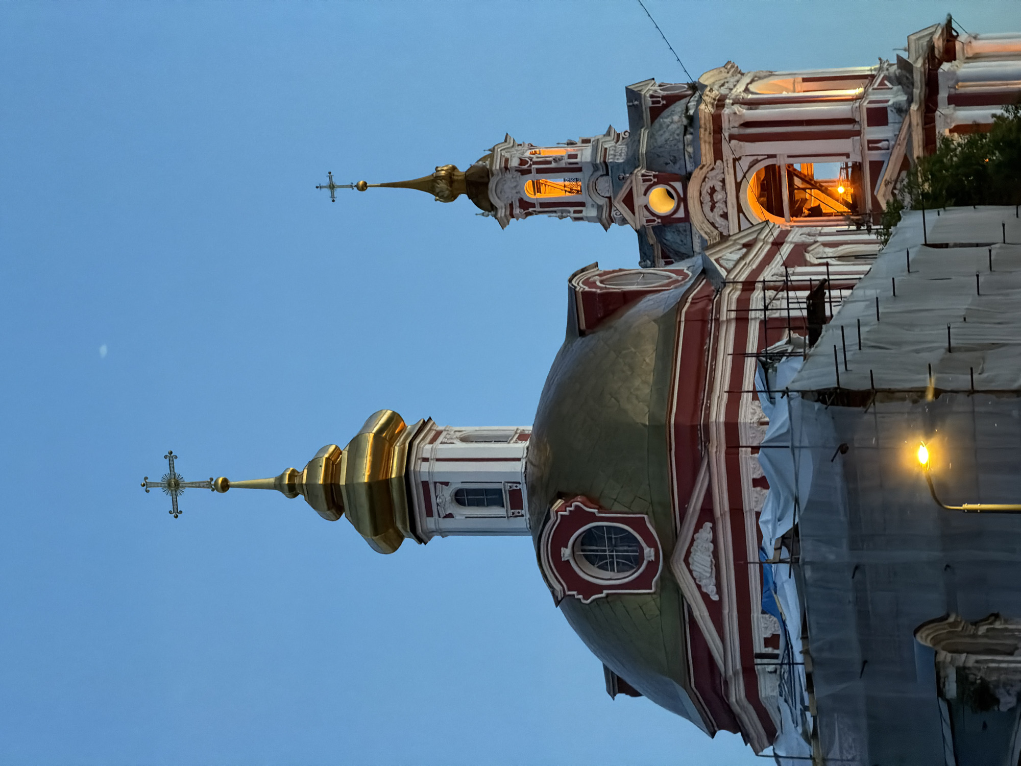 The image depicts the Church of the Archangel Gabriel, commonly known as the Menshikov Tower, located on Arkhangelsky Lane in Moscow, Russia, at dusk or early night.
In the foreground, two distinct church structures, adorned in red and white with gold domes, rise against a clear, deep twilight blue sky. The larger structure on the left features a broad, flattened golden dome. Below the dome, the building exhibits red and white striped walls with classical architectural details. An oval window (oculus) framed in red and white is prominent on the dome's base. Above the main dome, a white octagonal lantern with arched windows is capped by a smaller golden onion-shaped cupola, topped with a slender golden spire and an ornate Orthodox cross displaying a sunburst motif. The lower section of this structure is partially obscured by scaffolding covered in light-colored tarpaulin or netting, indicating ongoing renovation. A bright yellow street light illuminates a portion of the scaffolding and the base of the structure in the lower left.
To the right, a taller, more slender bell tower stands, sharing the red and white striped facade and classical detailing. Its upper tiers feature arched openings that reveal warm, golden artificial lighting from within. A circular opening, possibly a clock face, is visible below the illuminated archways, also emitting a soft yellow glow. This bell tower is also topped with a golden onion-shaped cupola, a slender golden spire, and an Orthodox cross. A faint, white crescent moon is visible in the upper left quadrant of the sky. No people are visible in the frame.
