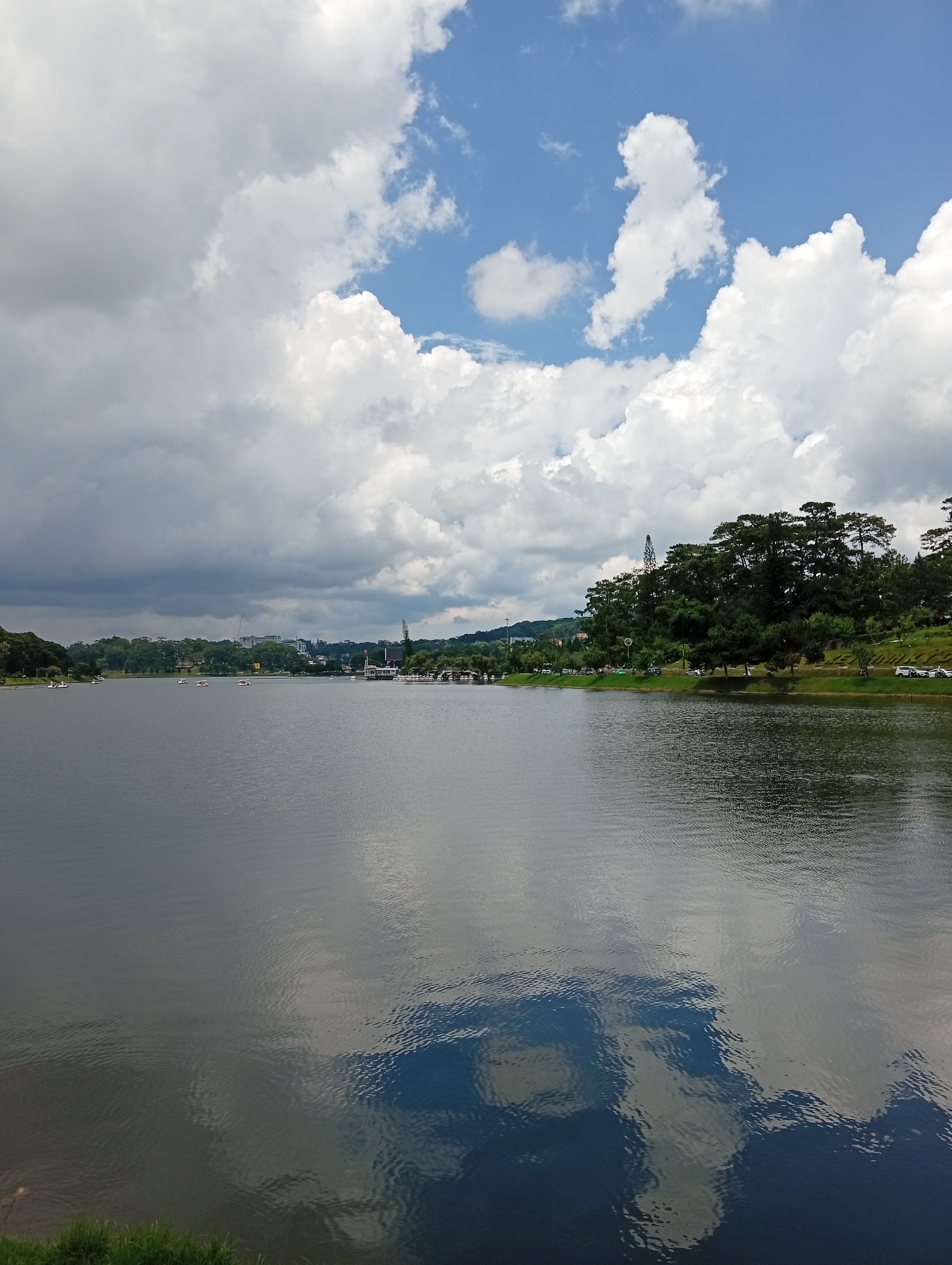 The image depicts Xuan Huong Lake in Da Lat, Vietnam. The foreground is dominated by the calm, dark water of the lake, reflecting the sky and surrounding scenery. The surface of the water exhibits subtle ripples, breaking up the reflections of the clouds and trees. The middle ground shows the far bank of the lake, characterized by a dense line of dark green pine trees. Beyond the trees, buildings and infrastructure of the city are visible, including some taller structures and a tower. A white building with a distinct architectural style is prominent on the right side of the far bank. Several small boats, some appearing to be pedal boats, are scattered across the lake, indicating recreational activity. The background consists of a sky filled with a mixture of white, fluffy cumulus clouds and darker, more ominous storm clouds, suggesting variable weather conditions. The overall impression is of a scenic natural landscape integrated with urban elements, typical of Da Lat.