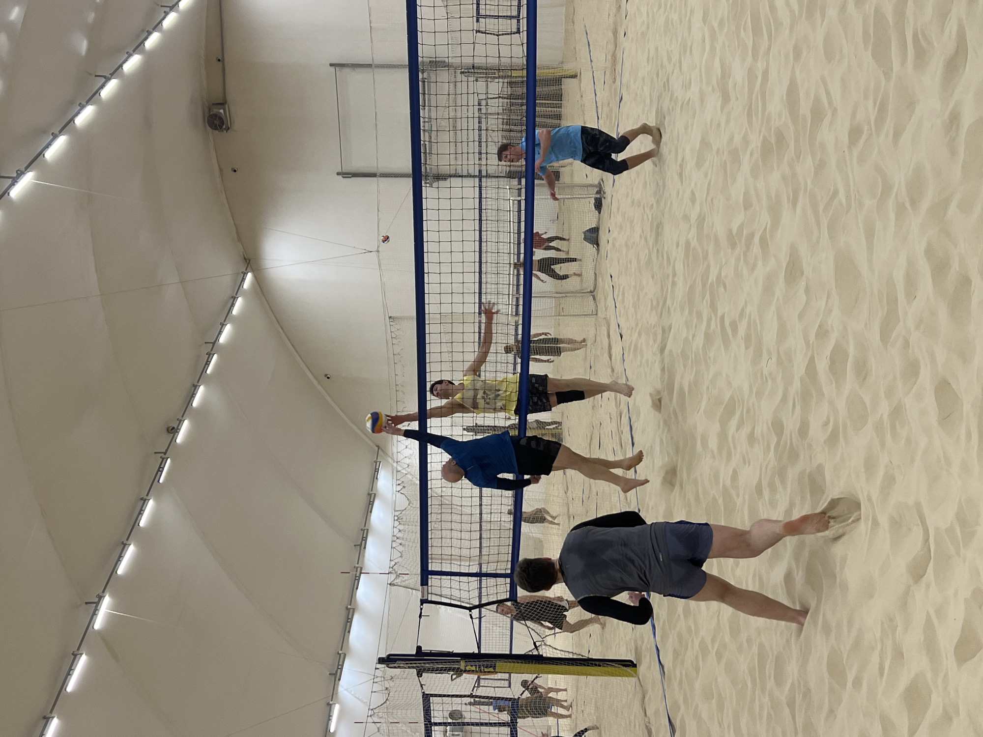 The image depicts a beach volleyball game being played indoors on a sand court. A volleyball net divides the court, with several players engaged in the game on both sides. One player in a blue shirt is spiking the ball. Other players are attempting to block the spike or are positioned to receive the ball. Spectators can be seen blurred in the background behind the net. The indoor facility has a high, curved ceiling with artificial lighting. It is not possible to identify the exact location within Saint Petersburg, Russia, from the given image.