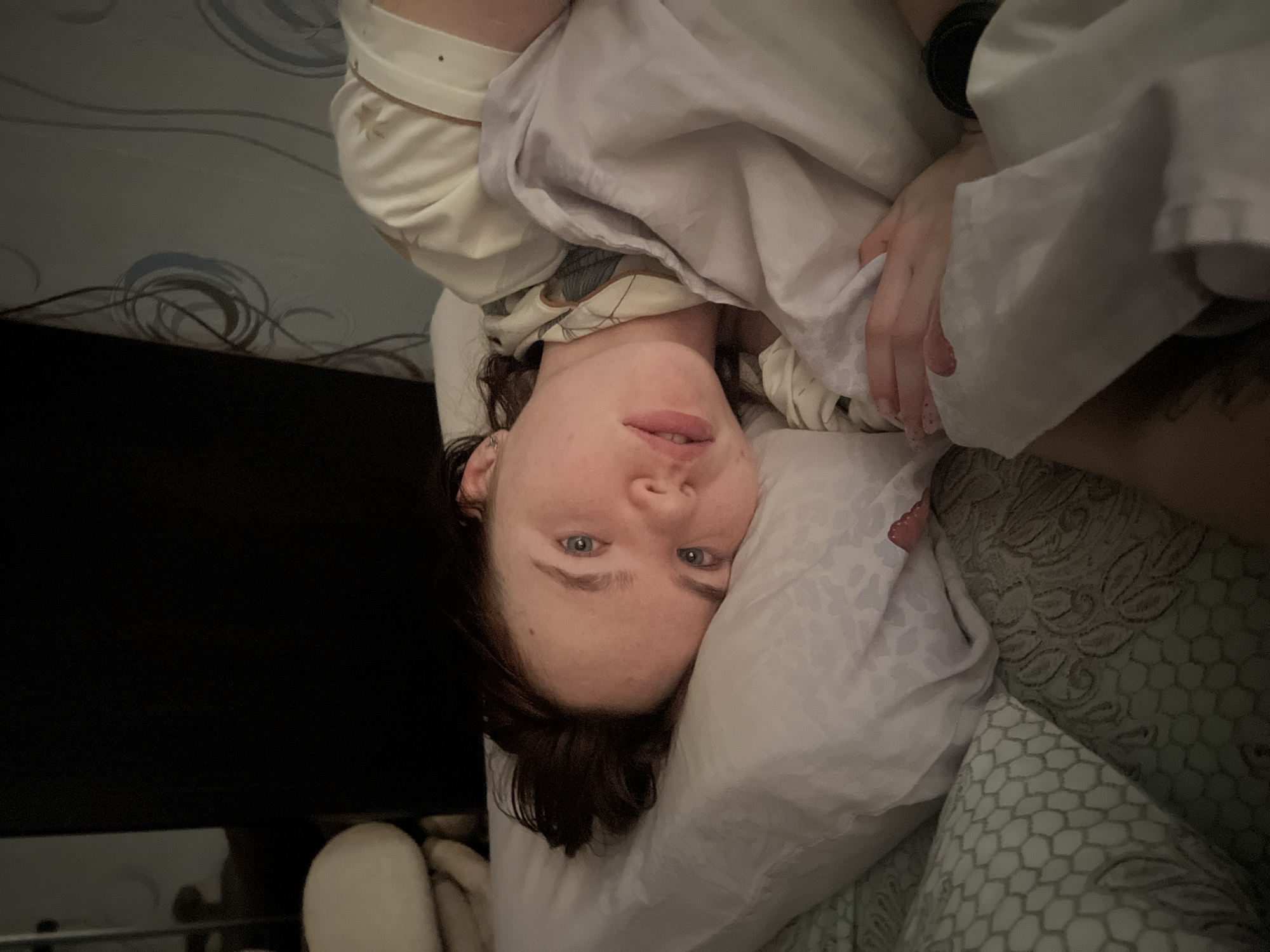 A Caucasian woman with short, dark hair is lying in bed, facing the camera. She has fair skin and blue eyes. She is partially covered by a white blanket or comforter and is resting her head on a white pillow. She appears to be wearing a light-colored pajama top. Her left wrist is visible, and there appears to be a dark-colored watch or bracelet on it. In the background to the left, there is a black object, likely a television, and wallpaper with a swirling design. The bedclothes include a patterned pillowcase or fitted sheet. The location is assumed to be an indoor domestic setting in Chaikovsky, Russia.
