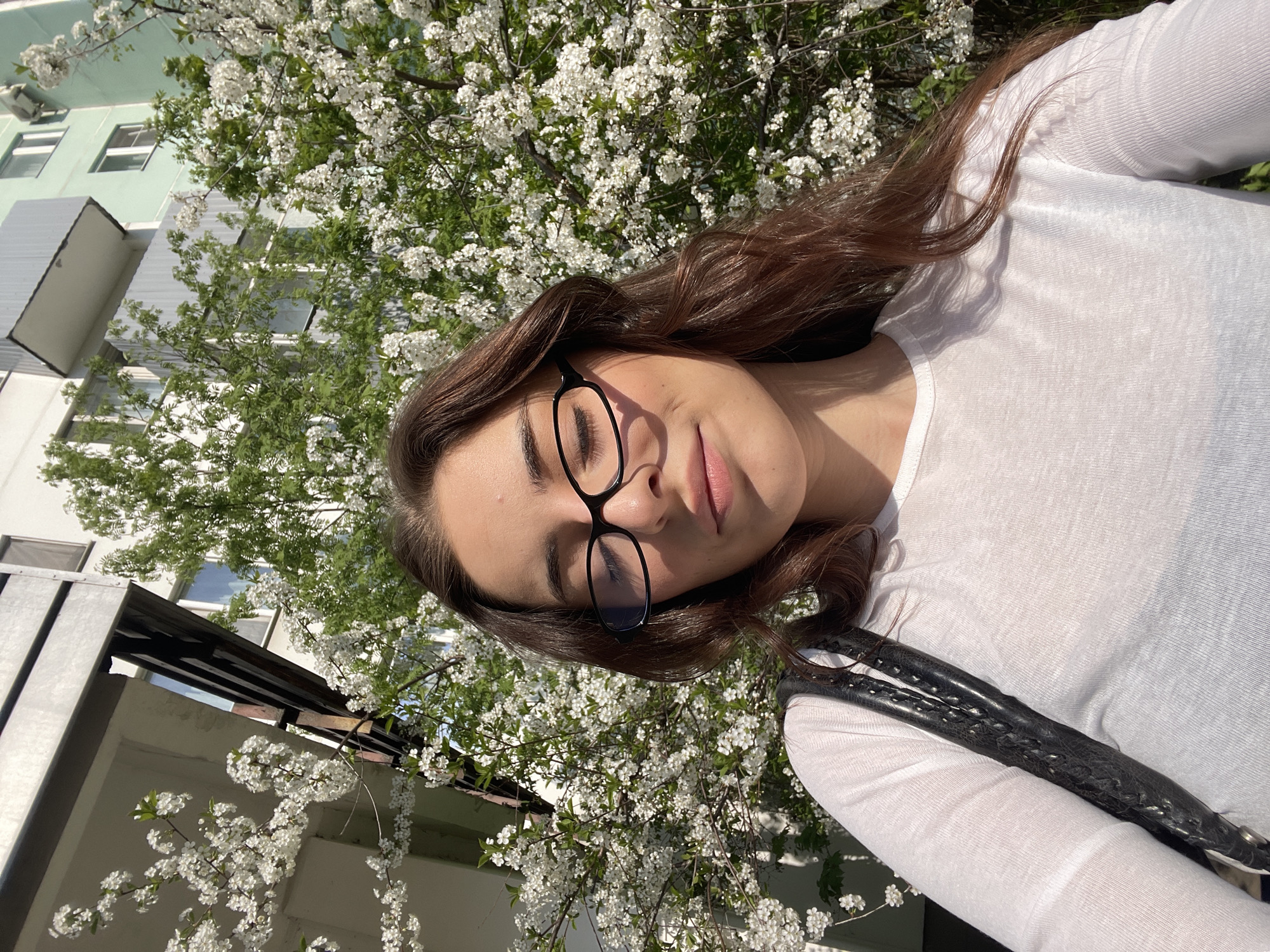 A young woman with long brown hair and glasses is posing for a selfie in Moscow, Russia. She is wearing a white long-sleeved shirt and has a black bag strap over her shoulder. The background consists of a building with white window frames and a tree with white blossoms. The angle of the shot is tilted, creating a disoriented perspective. The lighting suggests a sunny day.
