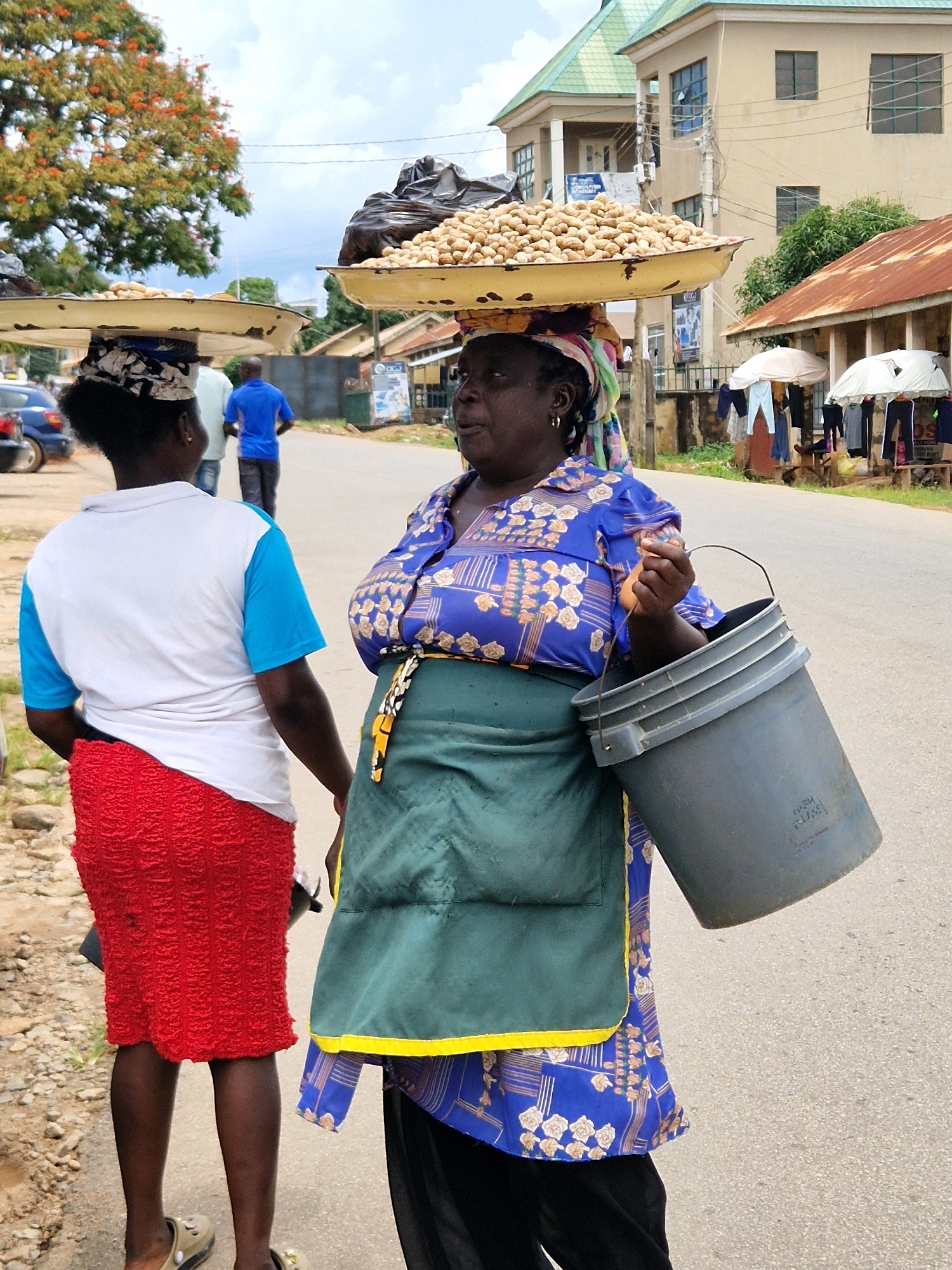 The image depicts a street scene in Jos, Nigeria, with two women carrying goods on their heads. The woman on the right, wearing a blue patterned blouse and a green apron, has a large tray of peanuts balanced on her head and is holding a grey bucket. The woman on the left, in a white and blue shirt and a red skirt, also has a tray on her head, though the contents are not clearly visible. In the background, a man in a blue shirt walks away from the camera. Buildings and trees line the street, with a clear sky overhead. The overall impression is of daily life and commerce in an urban Nigerian setting.