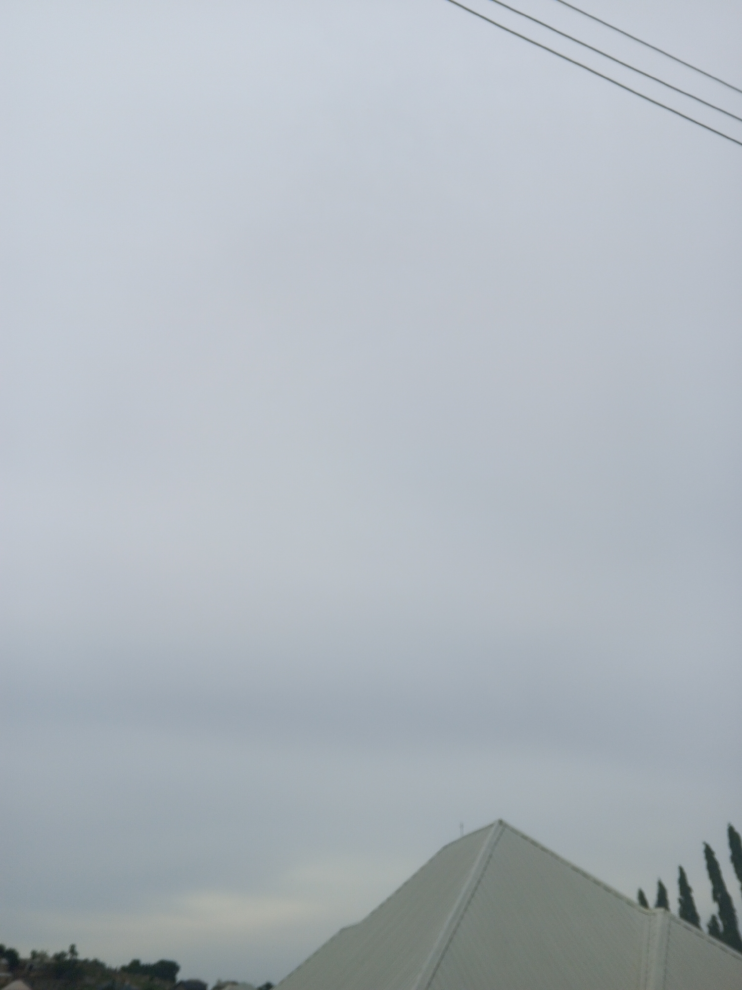 The image shows a cloudy, overcast sky above a portion of a white, angular building with a peaked roof. In the distance, there is a line of dark, conical trees and a hint of other structures. Three thin, dark lines, possibly power lines, are visible in the upper right quadrant of the sky. The scene appears to be an outdoor view of a modern structure with natural elements. The location is stated to be in the city of Ashata, Nigeria.