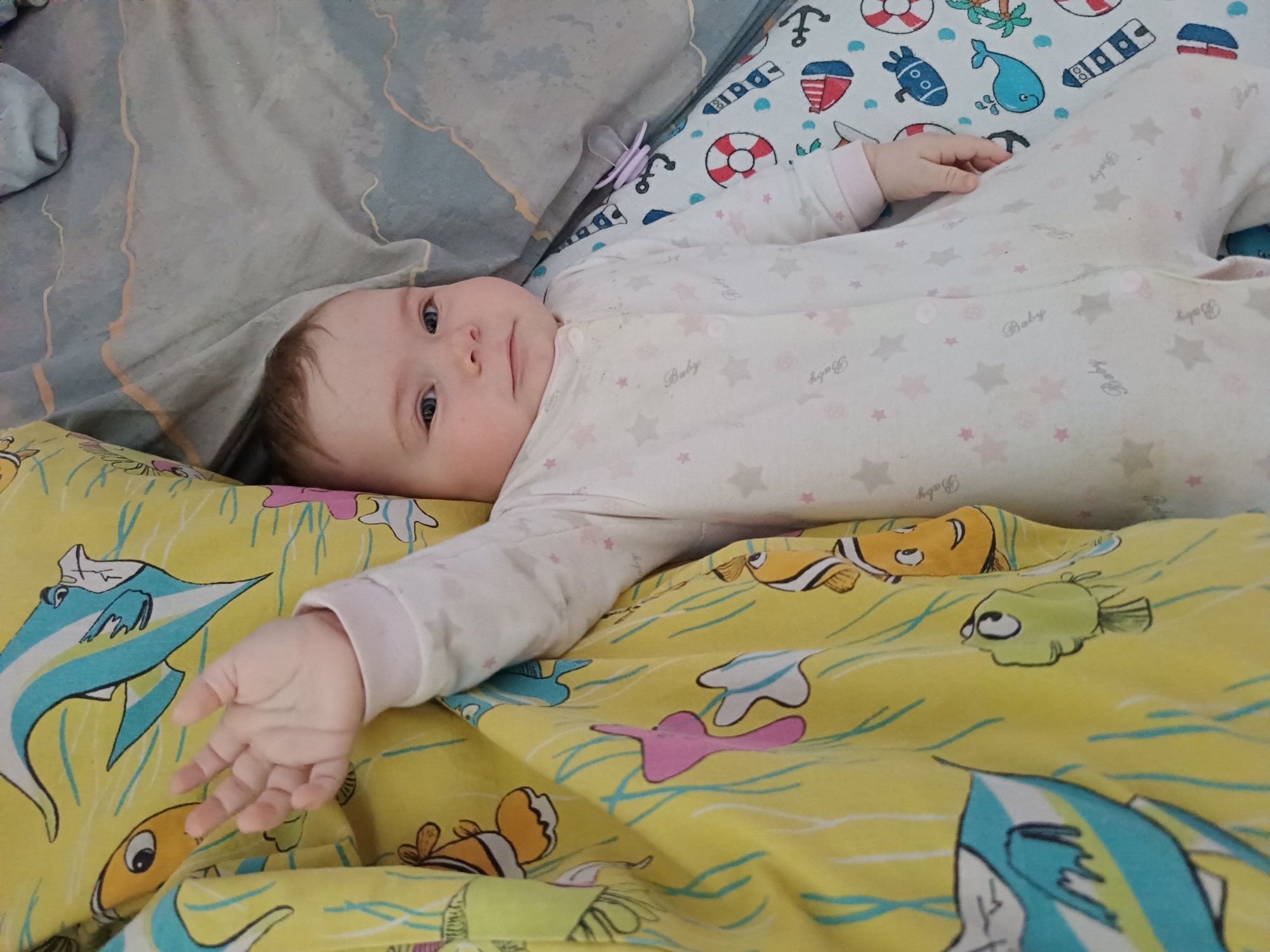 A baby lies on patterned bedding. The baby is wearing a white onesie with pink star and "Baby" patterns. The bedding features nautical-themed illustrations such as anchors, life preservers, and various sea creatures, as well as yellow bedding with illustrations of colorful fish. A purple pacifier rests nearby.
The location is not discernible from the image.
