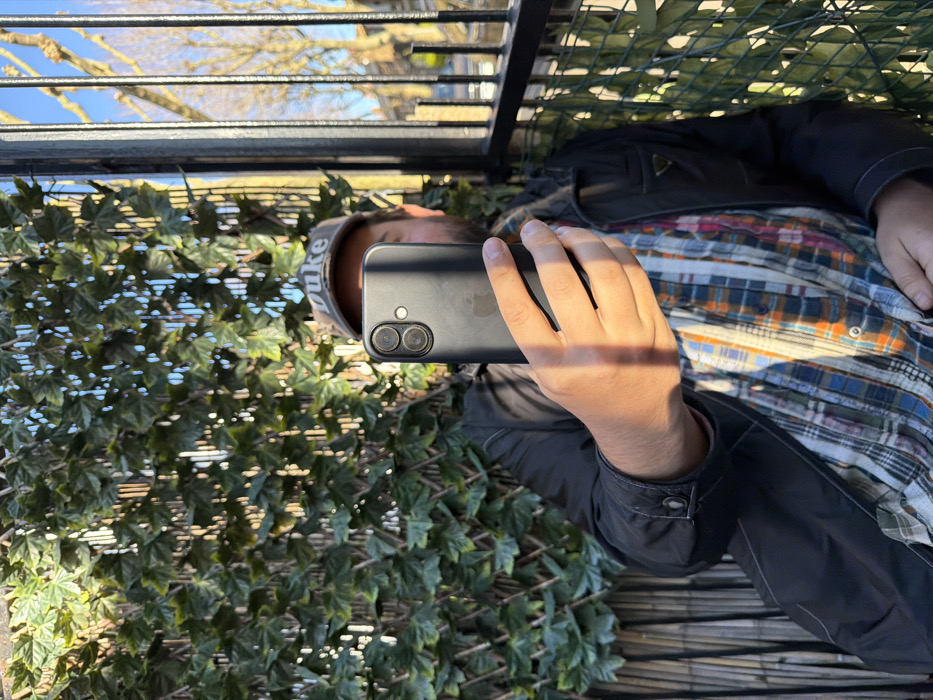 The image depicts a male individual, partially obscured by a dark-cased Apple iPhone held upright in his right hand (viewer's left), positioned as if taking a photograph or selfie. He wears a beige "DUKE" baseball cap, a dark-colored jacket with a small triangular emblem on the left chest pocket area, and a multi-colored plaid shirt with orange, blue, white, and red tones underneath. The scene is an outdoor urban environment, consistent with the city of London, United Kingdom, observed under strong direct sunlight and a clear blue sky. To the left and behind the individual, a lattice-like structure is covered in artificial green ivy. To the right, a black metal railing extends vertically, beyond which bare deciduous trees and indistinct residential or commercial buildings line what appears to be a street or open area. Direct sunlight casts distinct shadows across the individual's hand and arm, as well as from the metal railing. The rear of the iPhone, including its dual-camera system and Apple logo, is visible.
