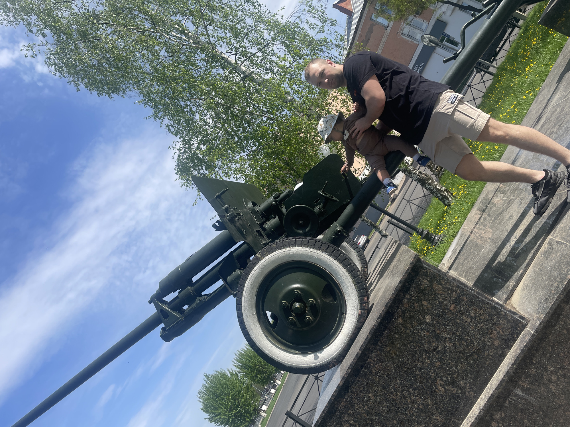 A low-angle, sunny outdoor shot depicts a man and a toddler interacting with an artillery gun on display. The gun is dark green with a long barrel and white-walled tires, mounted on a dark granite base. The man, dressed in a black t-shirt and khaki shorts, leans over the gun, apparently assisting the toddler, who is wearing a hat and brown outfit. A building is visible in the background. The image was likely taken in Votkinsk, Russia, as the artillery gun is a local monument.