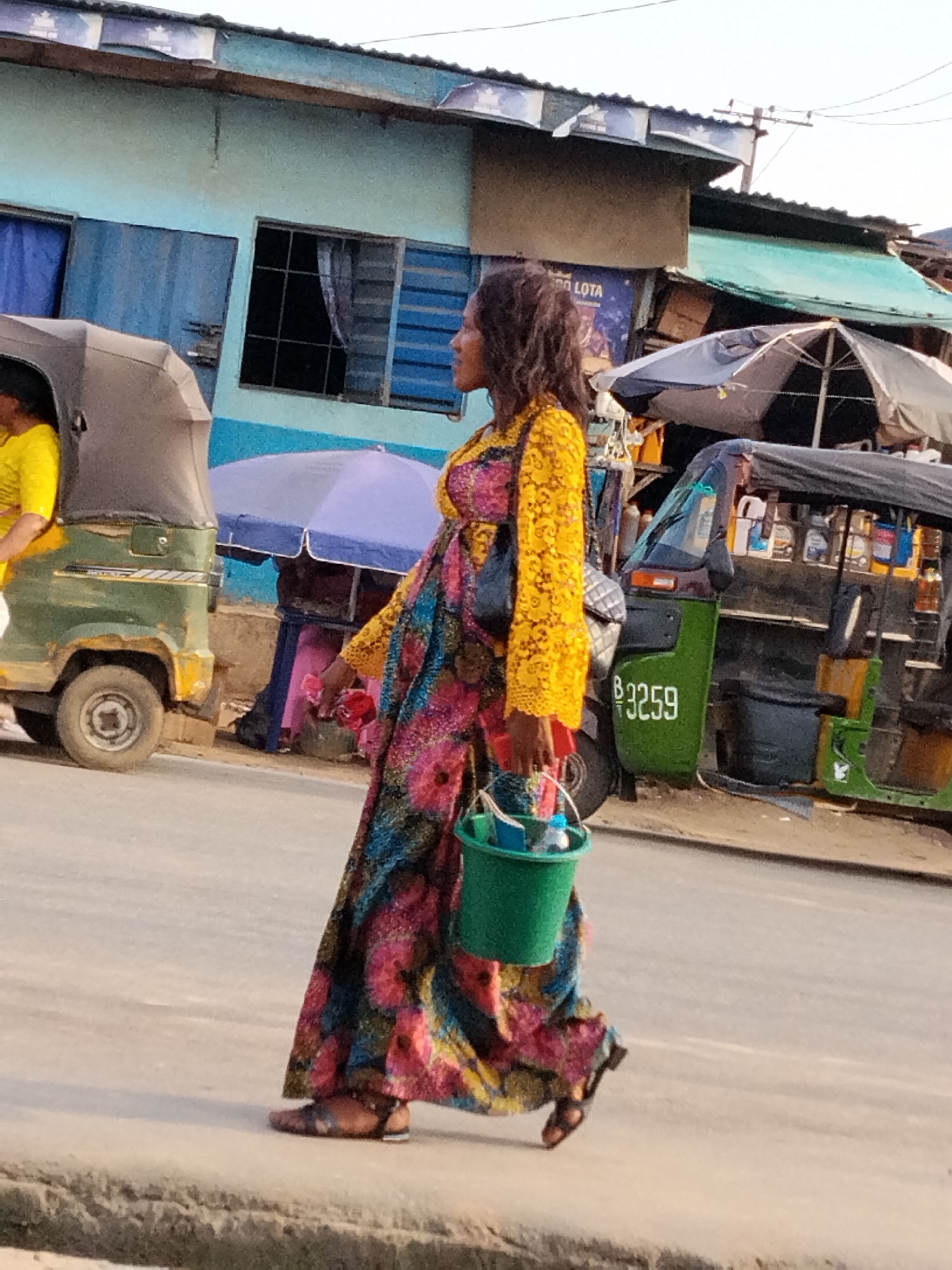 A woman walks along a paved road in Aba, Nigeria. She is wearing a long, patterned dress with a yellow lace top and carrying a green bucket filled with blue and white objects. In the background, there are parked auto-rickshaws, one of which has the number "B 3259" visible on its side. Buildings with a mix of blue and green exteriors are also present, along with market stalls and umbrellas.
