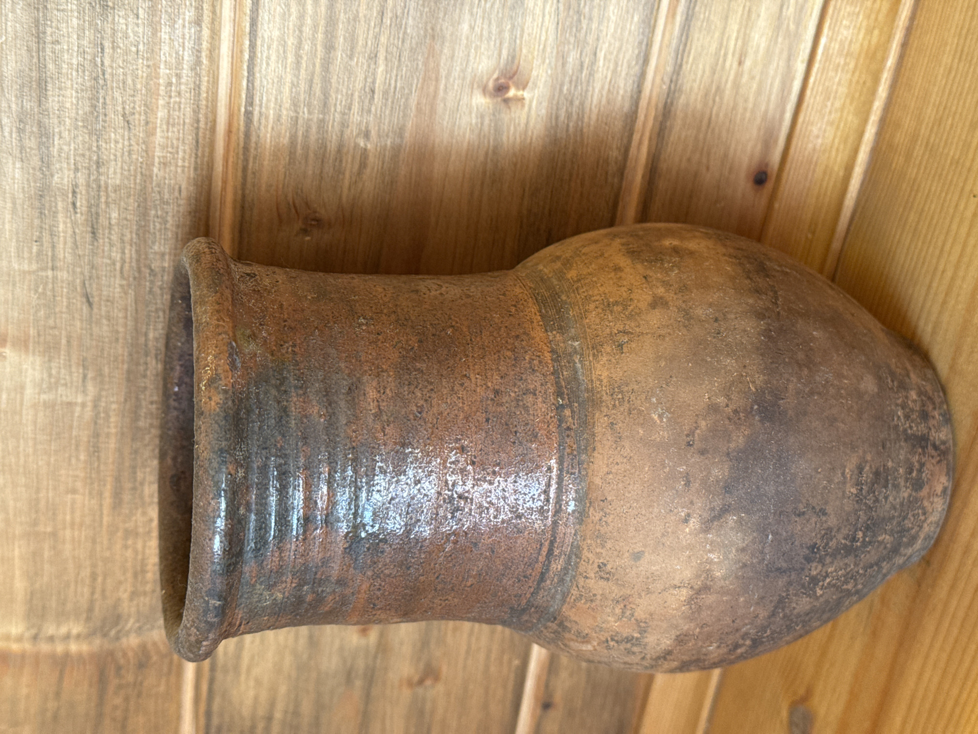 An earthen pot lies on its side against a wood plank wall. The pot has a wide mouth and a rounded body with a tapering base. The surface of the pot is mottled with shades of brown and grey. The wood wall is light brown. The location is not in the image.