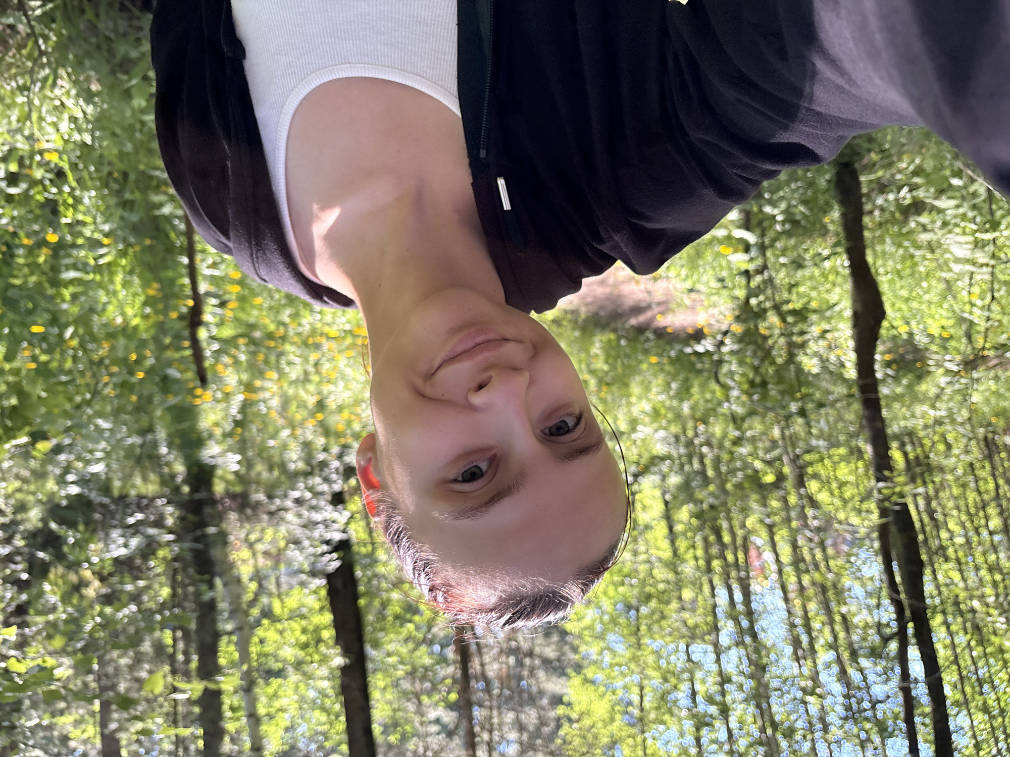 A young woman with auburn hair pulled back is taking a selfie outdoors. She is wearing a white tank top and a black zip-up hoodie. She is smiling gently at the camera, with a slight tilt of her head. The background is a sun-dappled forest with many trees and green foliage. Yellow wildflowers are scattered throughout the undergrowth. The lighting suggests it is daytime. The location appears to be a natural park or wooded area, consistent with areas within or near Chaikovsky, Russia, a city known for its surrounding natural beauty and forests.