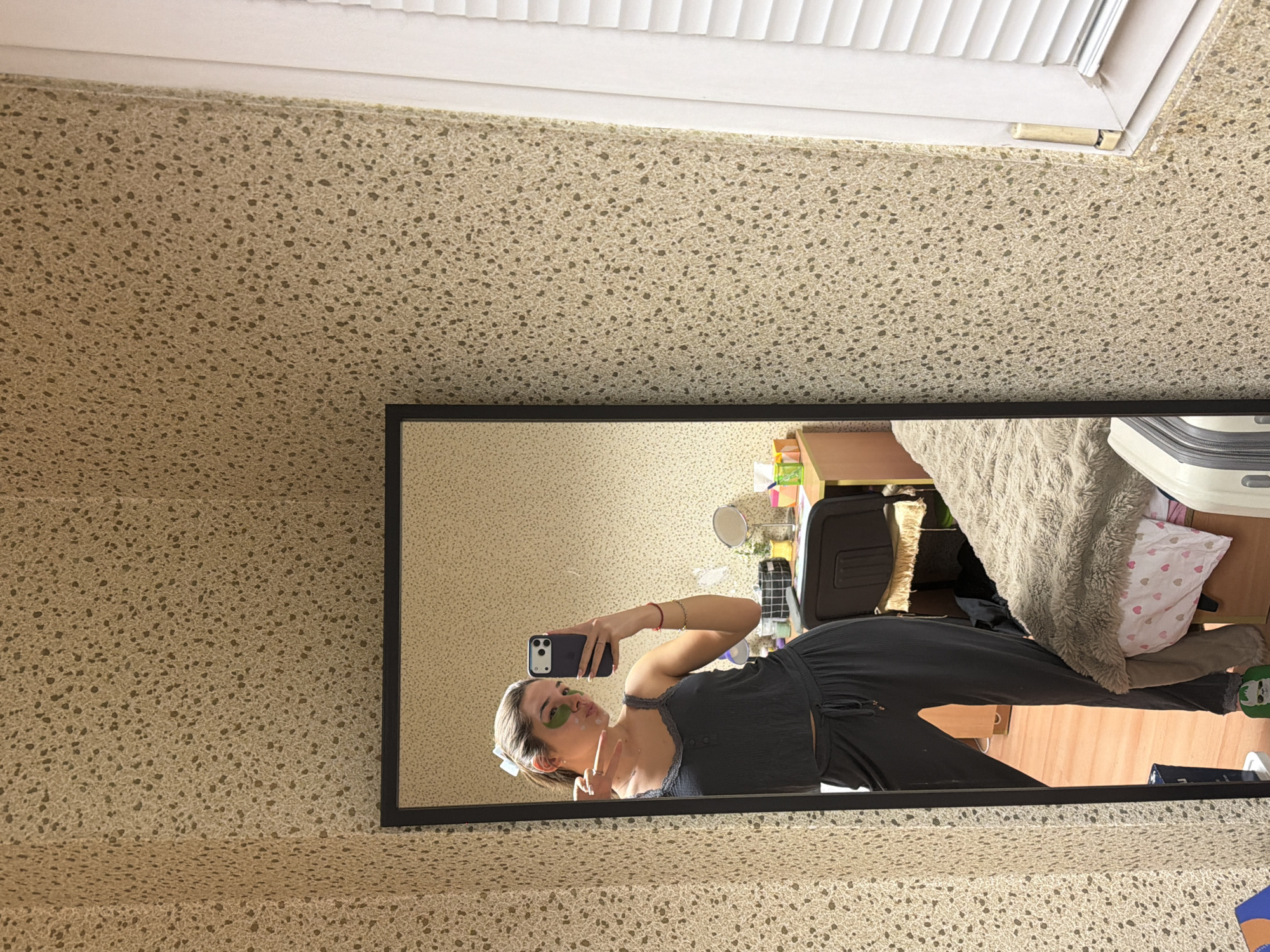 The scene is an indoor domestic environment, stated to be located in Sofia, Bulgaria. A young adult female is depicted via her reflection in a black-framed, floor-length mirror, taking a selfie. She has light-colored hair pulled back into a ponytail, secured with a light-colored bow. Her face displays green under-eye patches and a white facial product dabbed on her right cheek and chin, beneath dark-rimmed glasses. She is dressed in a dark gray ribbed camisole with lace trim and matching drawstring lounge pants. She holds a dark-colored smartphone with a light-colored case in her right hand, aimed at the mirror. Her left hand is raised, forming a 'peace' sign. On her right wrist, a thin red string and a gold-colored chain bracelet are visible. A green slipper is partially visible on her left foot.
The room features light beige wallpaper with a dense, speckled pattern covering at least two walls. Reflected behind the subject are various elements: a light-colored wooden desk cluttered with personal care items, including two circular mirrors, and a dark office-style chair draped with light fabric. To the right of the desk, a bed is covered with a thick, textured light brown blanket over a white sheet patterned with pink hearts. A light-colored suitcase is partially visible at the foot of the bed. The flooring visible in the reflection is light-colored wood. To the far right of the main image, a white-framed window with white louvered blinds is partially visible.