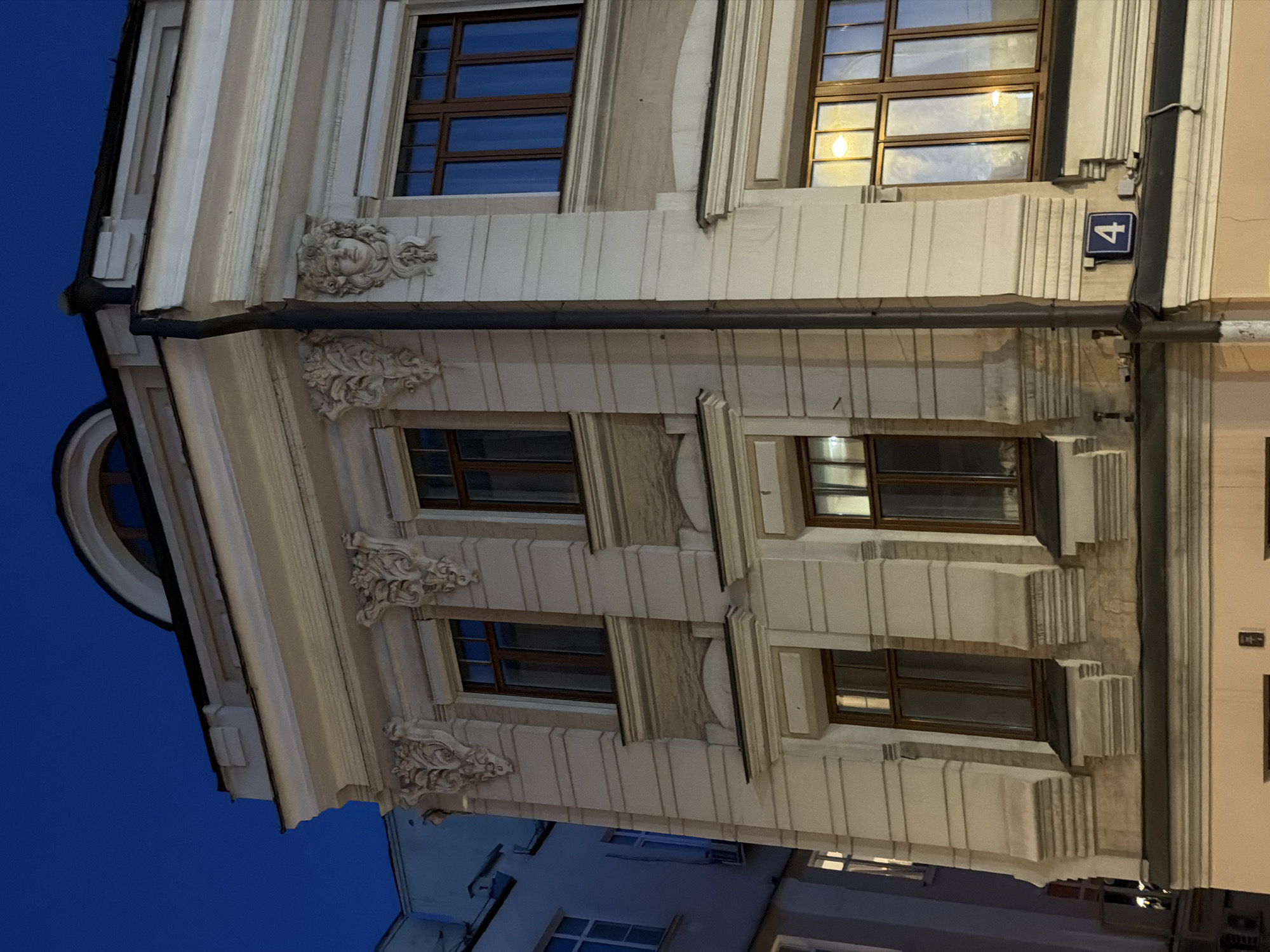 A low-angle shot shows the facade of a building, likely at night due to the dark blue sky in the background and artificial lighting illuminating the structure. The building has a beige or light gray color. It features several windows with dark frames. The architectural style is elaborate, with decorative elements such as carved ornaments, including what appears to be a sculpted face near the top left. A dark, thick pipe runs horizontally across the facade. A blue sign with the number "4" is visible on the right.