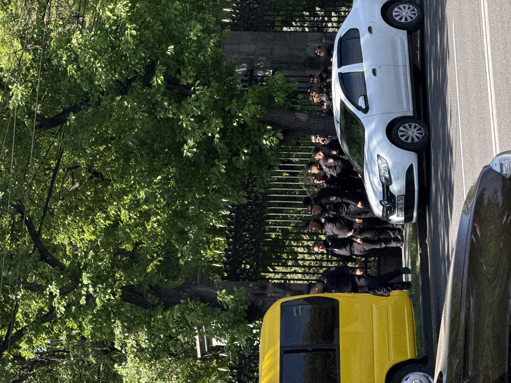 The image depicts a street scene in Yerevan, Armenia. A white car and a yellow van are parked on the right side of the road, facing the same direction. A group of uniformed individuals, possibly police officers, are standing behind a black iron fence that lines the sidewalk. A tree with dense green foliage is to the left of the fence and the group of officers. The lighting suggests it is daytime.