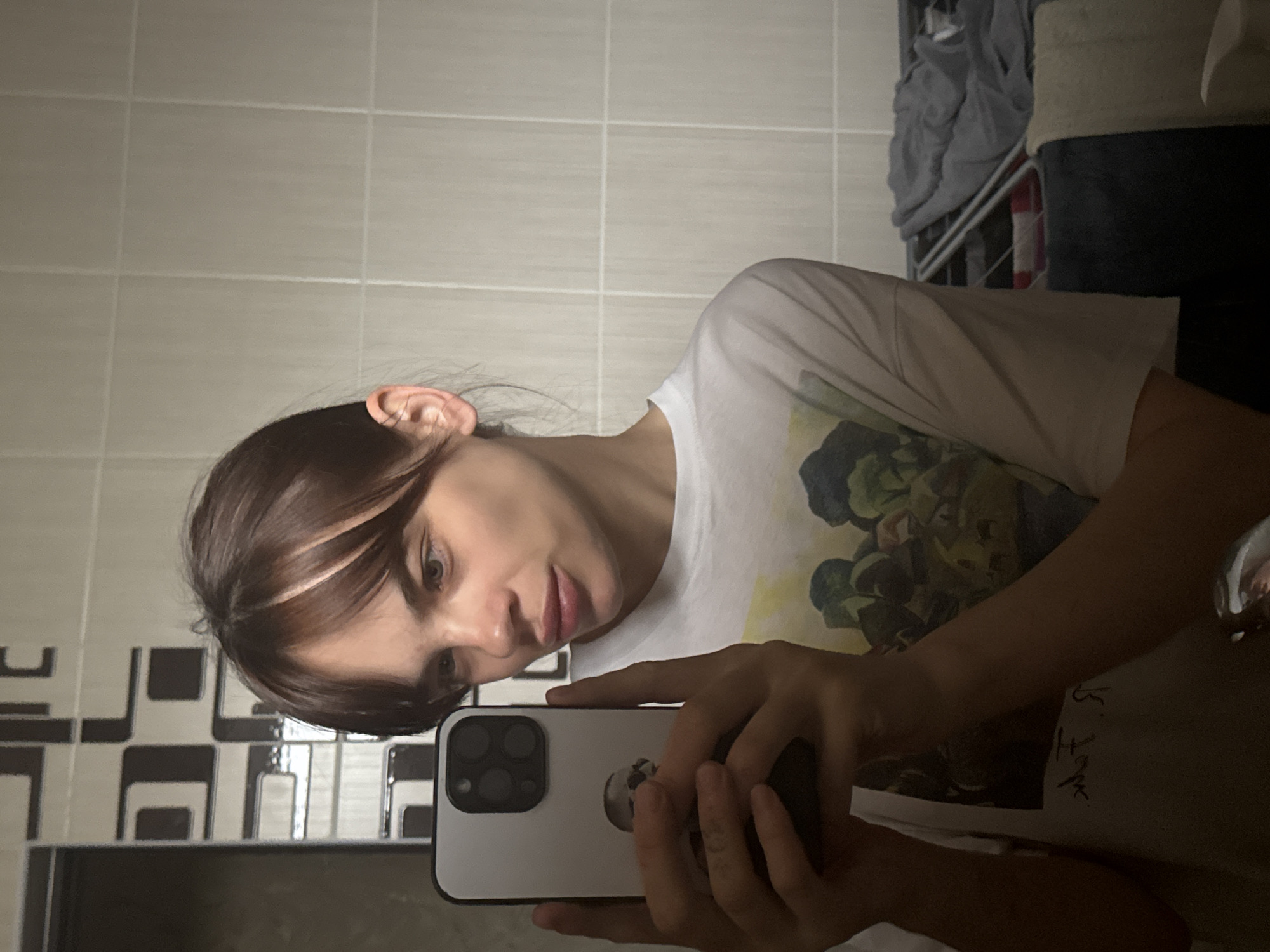 A woman is taking a selfie in a bathroom. The bathroom has white and gray tiled walls. She is holding a white iPhone with multiple camera lenses. The woman has brown hair with a blonde streak near her forehead. She is wearing a white t-shirt with a dark-colored graphic. The background includes a laundry basket with clothes. It is unclear whether the location is in Rostov-on-Don, Russia.