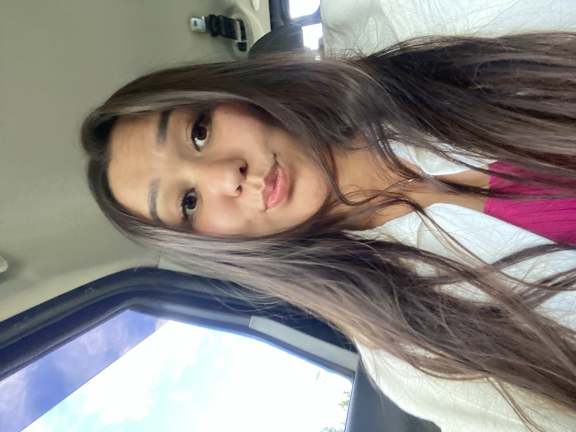 A young woman with long, dark brown hair is taking a selfie inside a vehicle. She is looking at the camera with her lips puckered. She is wearing a white jacket over a pink top. The background shows a car interior with a window to her left, revealing a blue sky with some clouds and trees outside. The image appears to be taken during the daytime. The location is not specified in Zilair, Russia, as this detail cannot be determined from the visual information provided.