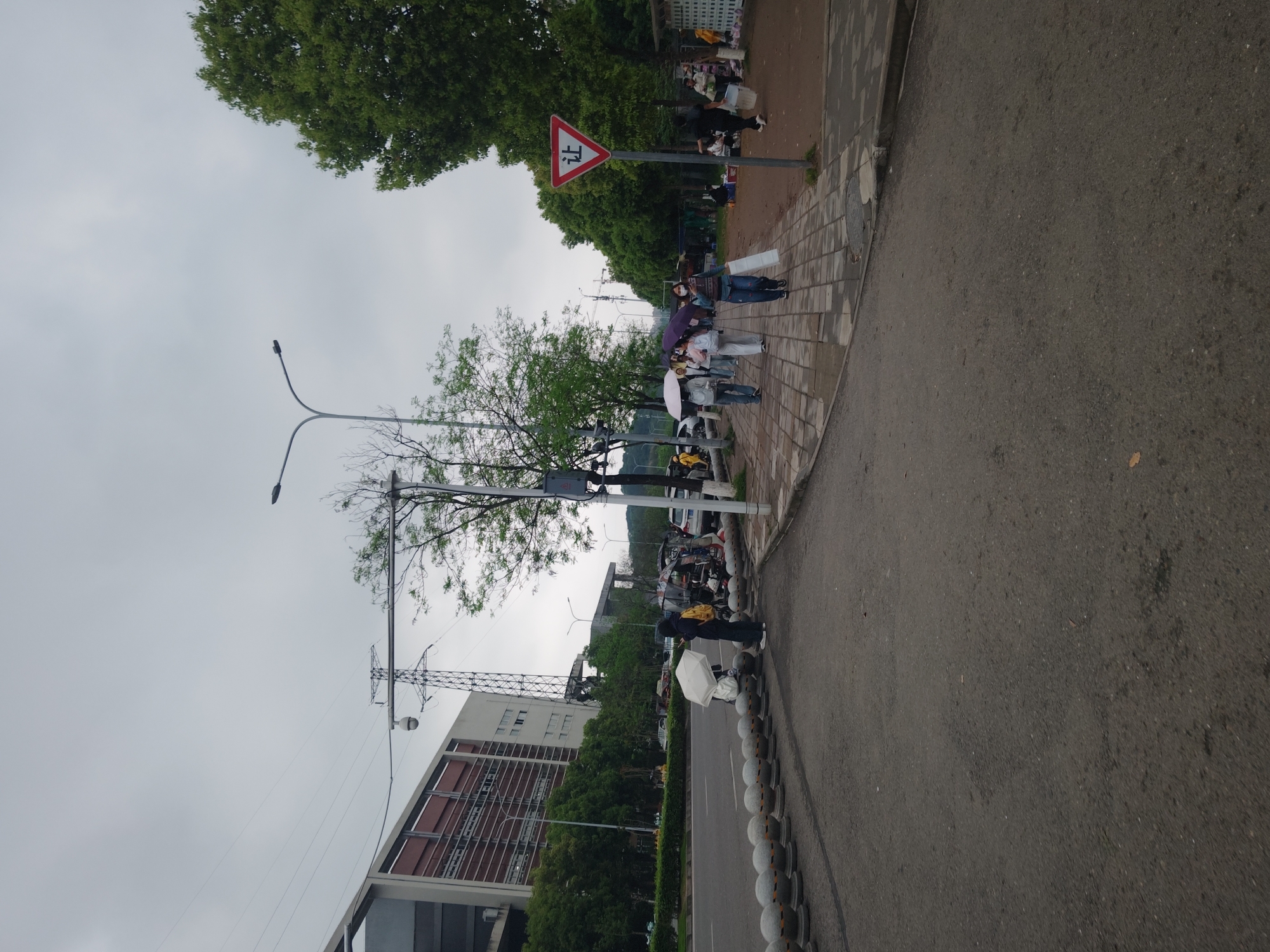 The image depicts a street scene in the Shapingba District of China on a cloudy day. A paved road occupies the right side of the image. A sidewalk runs parallel to the road, lined with a row of spherical concrete bollards. Several people are walking along the sidewalk, some carrying umbrellas. A red triangular sign with Chinese characters is visible, indicating a pedestrian crossing. Lampposts are seen extending into the sky. A building with a multi-story facade is visible in the background, along with greenery consisting of trees and shrubs. Overhead electrical power lines are present.