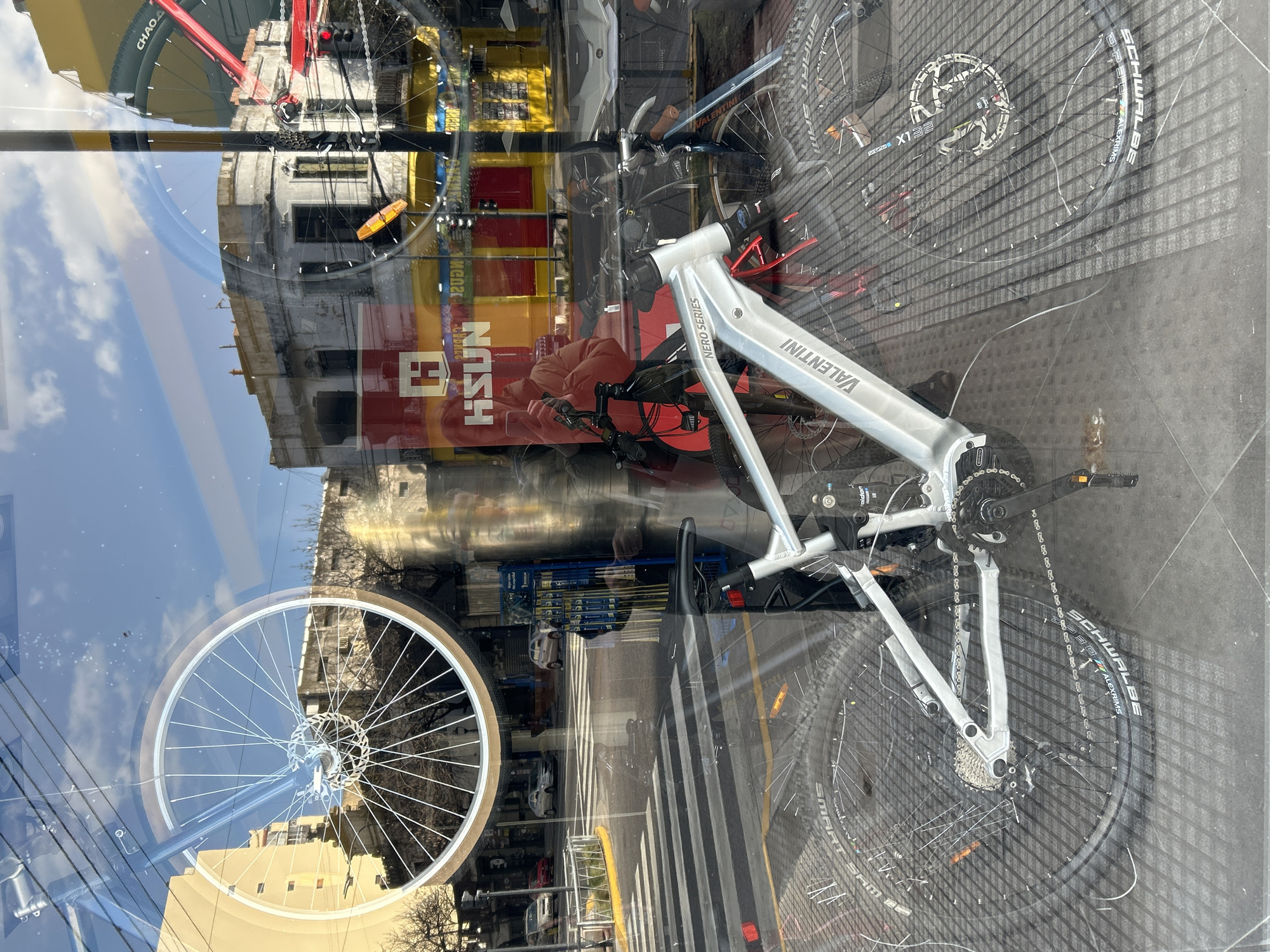 The image is a reflection of a street scene in Buenos Aires, Argentina, captured through a shop window. A silver electric bicycle, identified by "VALENTINI NERO SERIES" text on its frame, is prominently displayed inside the shop and is the main subject. The bicycle is positioned at an angle, with its front wheel partially visible on the left and its rear wheel and drivetrain on the right. The shop's interior is also reflected, showing racks of what appear to be bicycle tires, and a red traffic light.
The reflection shows a street with a pedestrian crossing, cars, and buildings typical of Buenos Aires architecture, including a building with a yellow facade. The sky is blue with scattered clouds. A person is visible in the reflection, holding a phone and appearing to take a photo of the bicycle display. The overall lighting suggests it is daytime. The image is characterized by reflections and glare from the shop window, which overlay and distort the scene.