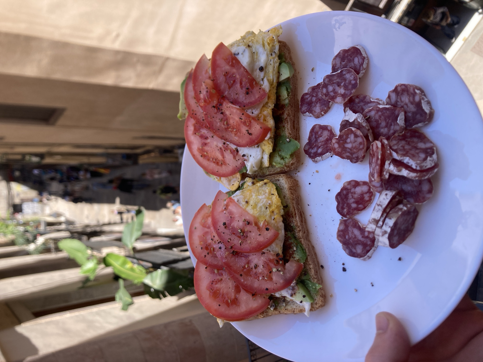 The image shows a white plate held in someone's hand, featuring two open-faced sandwiches and sliced salami. Each sandwich consists of a slice of toast topped with avocado, a fried egg, and tomato slices, sprinkled with black pepper. The salami is sliced into small, circular pieces and arranged on the right side of the plate. The background reveals a cityscape, with architectural features suggesting the location is Barcelona, Spain. Specifically, the buildings are likely on Rambla de Catalunya, as can be inferred from the balcony structure and planting visible in the image. The scene suggests an outdoor setting, possibly a balcony or terrace.