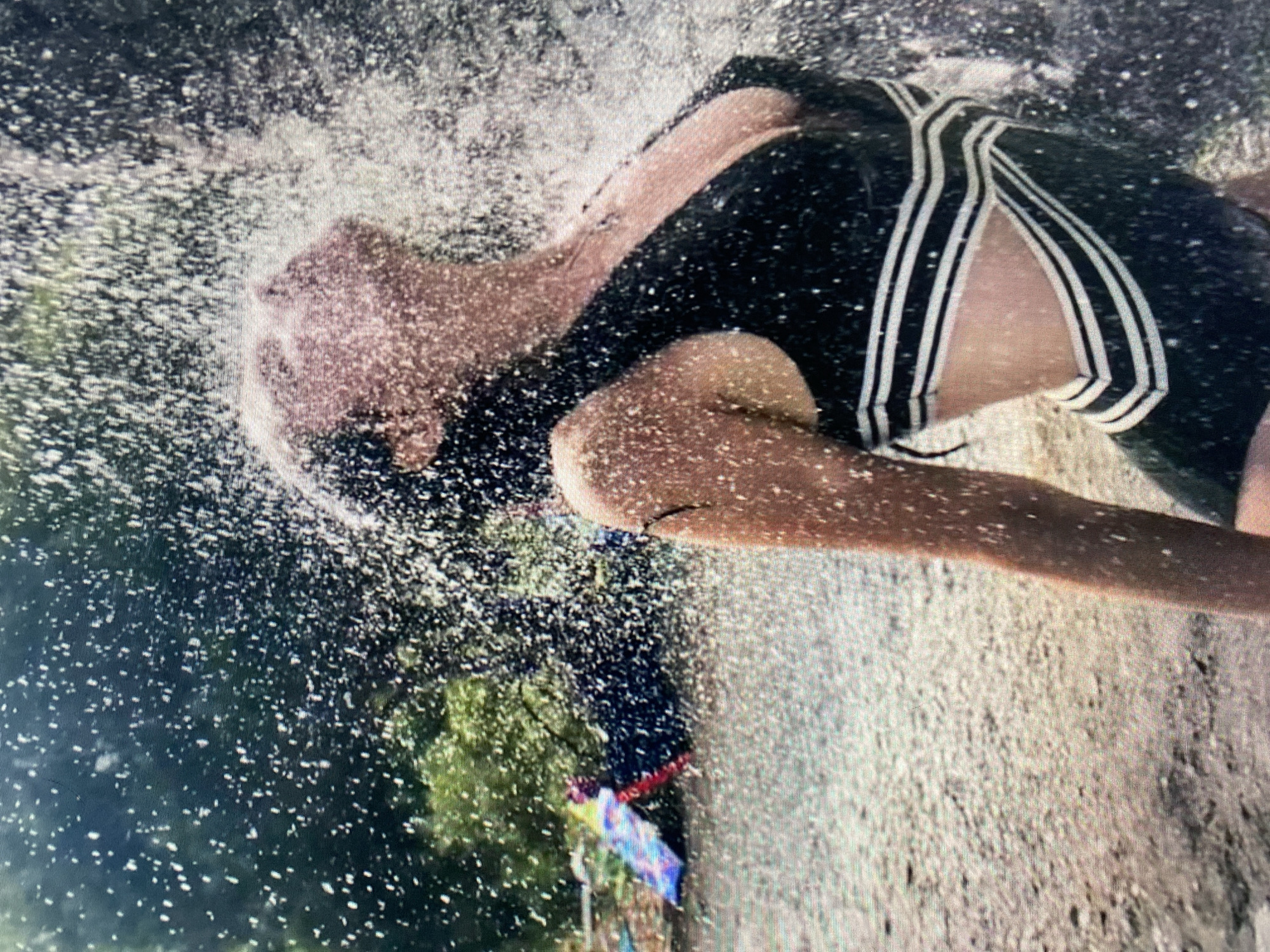 A woman stands outdoors, her head thrown back as water cascades over her face and body. She wears a black swimsuit with white stripes on the back. The backdrop consists of greenery, a concrete surface, and what appears to be a sprinkler creating the water spray. The location is Łódź, Poland.