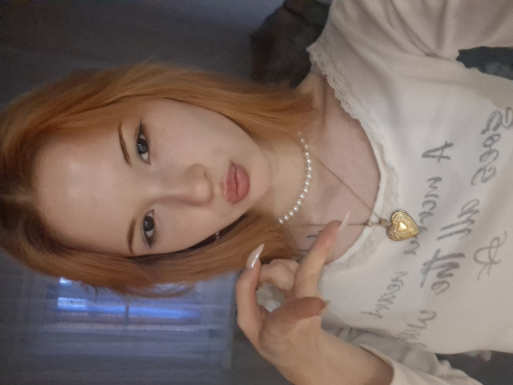 A young person with reddish-blonde hair and light skin is shown from the shoulders up. They are wearing a white top with lace trim at the neckline and writing on the front. A pearl necklace and a heart-shaped pendant hang around their neck. Their right hand is raised, forming a circle with their thumb and forefinger. The background is blurred, revealing a window with blinds and a hint of blue and gray tones. The location is Unknown, Unknown.