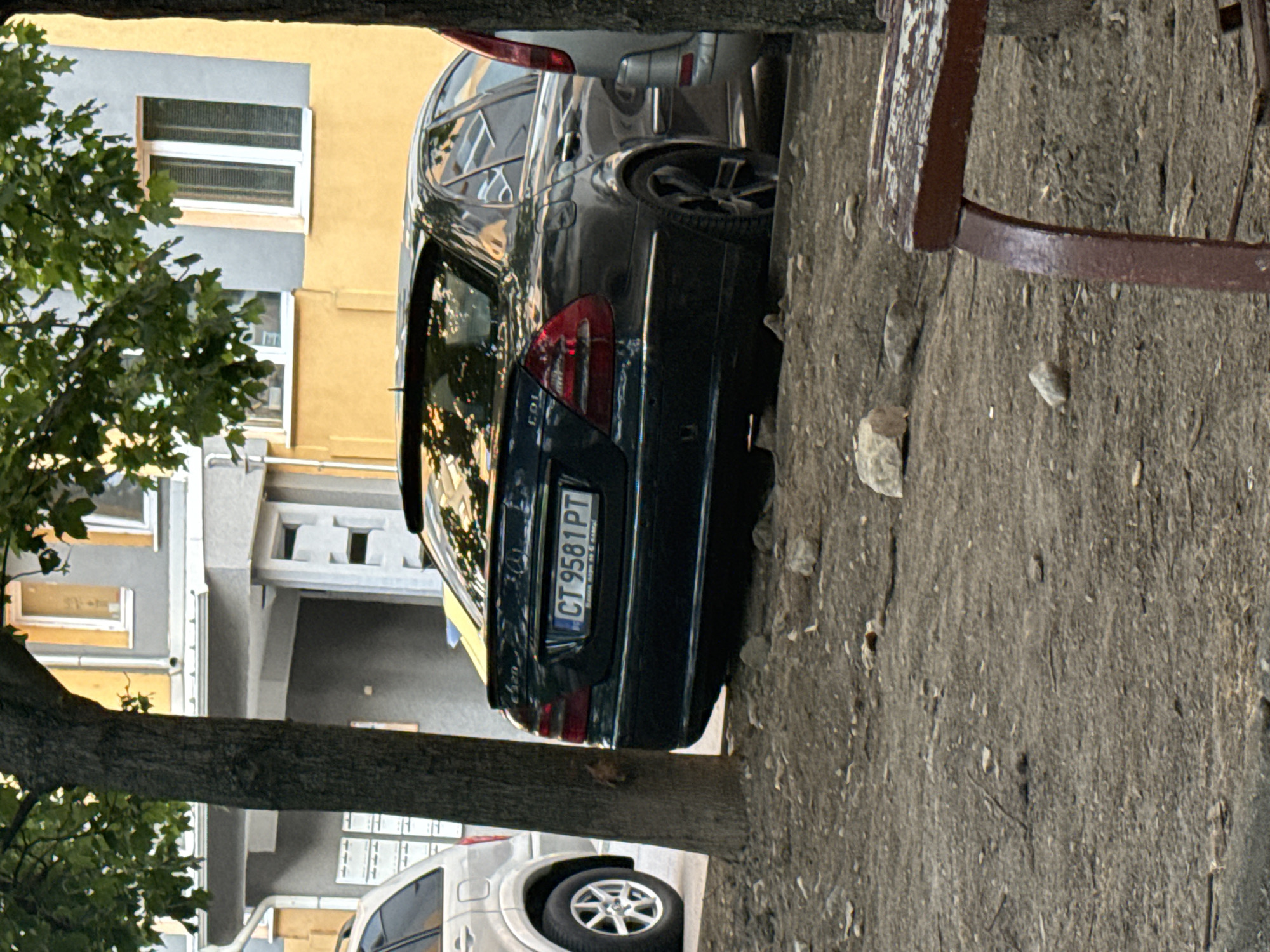 The image depicts a street scene in Stara Zagora, Bulgaria, with a dark-colored Mercedes-Benz sedan parked on the right side of the frame. The car has a license plate reading "CT 9581 PT". It is parked on a dirt and gravel area, partially obscured by a tree trunk on the left. In the background, a building with yellow and gray sections and various windows is visible. Another vehicle, a silver SUV, is parked to the left of the Mercedes. The foreground shows loose dirt, stones, and a portion of a wooden bench. The overall impression is one of an urban setting with vehicles and a building.