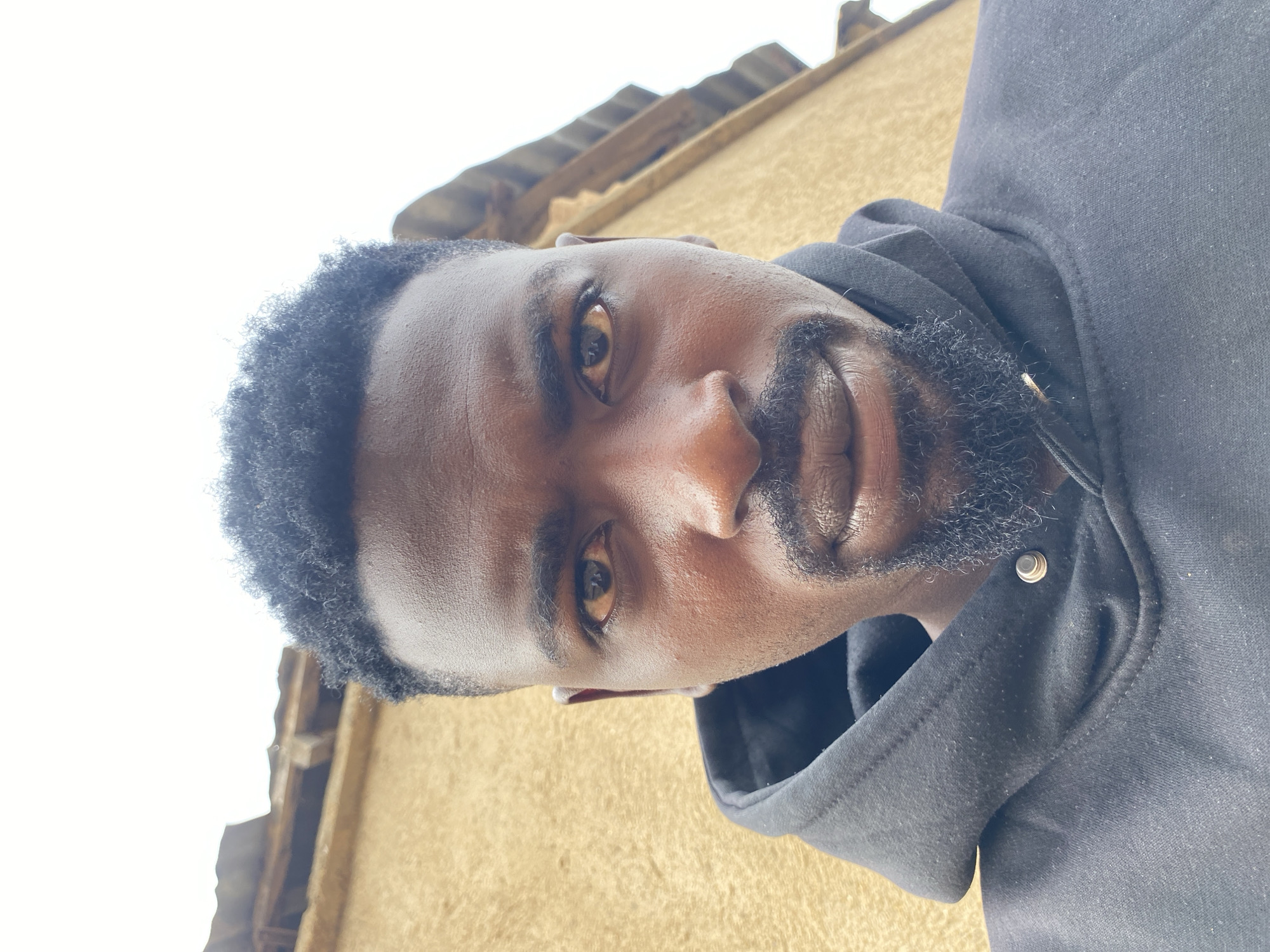 The image shows a dark-skinned man with short, dark, curly hair and a beard. He is wearing a black hoodie. The background shows a beige wall and a roof structure, suggesting an outdoor location. The photo was likely taken from a low angle, looking up at the man. Given the stated location, it is presumed to be Barakin Akawo, Nigeria.