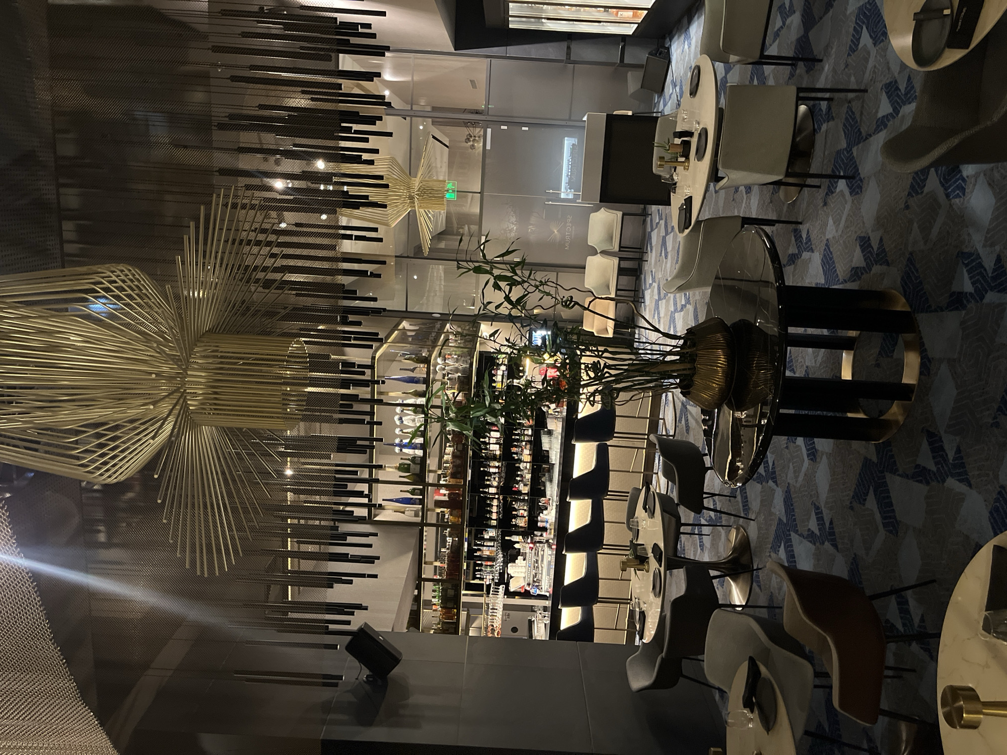 The image depicts the interior of a bar or restaurant, possibly in Sofia, Bulgaria. The scene features a modern aesthetic with a prominent display of alcoholic beverages on illuminated shelves behind the bar. There are several tables and chairs visible, some round and some with a more traditional design. Above, decorative lighting fixtures create an ambiance within the space. The floor appears to be patterned. Additionally, a green plant sits on the bar's counter.