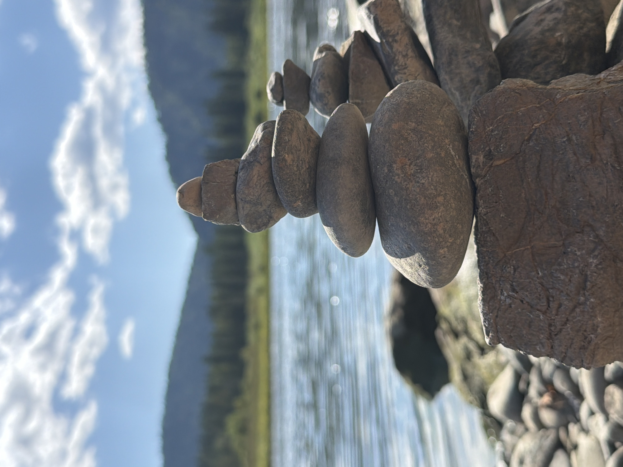 The image captures a close-up view of stacked rocks, or cairns, on the shore of a body of water, likely a river given the presence of a treeline and hills in the background. The water is calm and reflects the sky, which is partly cloudy with a bright blue expanse. The foreground is dominated by the carefully balanced stones, with some larger, rougher rocks forming the base. A second, smaller stack of stones is visible further back on the right. The environment appears natural and tranquil, with lush green vegetation and distant, forested hills under a daytime sky. The city of Ust-Kabirza, Russia, is not depicted or implied in this image.