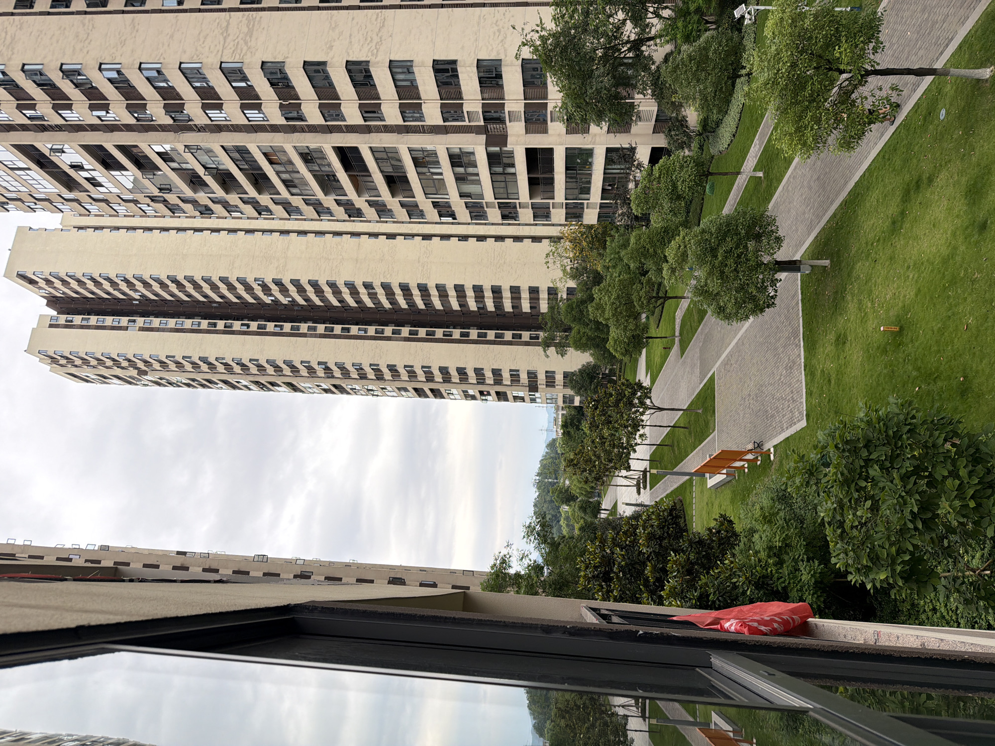 The image, taken from a high-angle view, shows a residential area in Chongqing High-tech Zone, China. Two tall apartment buildings dominate the scene, rising into a cloudy sky. Between the buildings is a landscaped park with green lawns, trees, and paved walkways with benches. The foreground shows a window frame with reflections and a red object on the ledge.