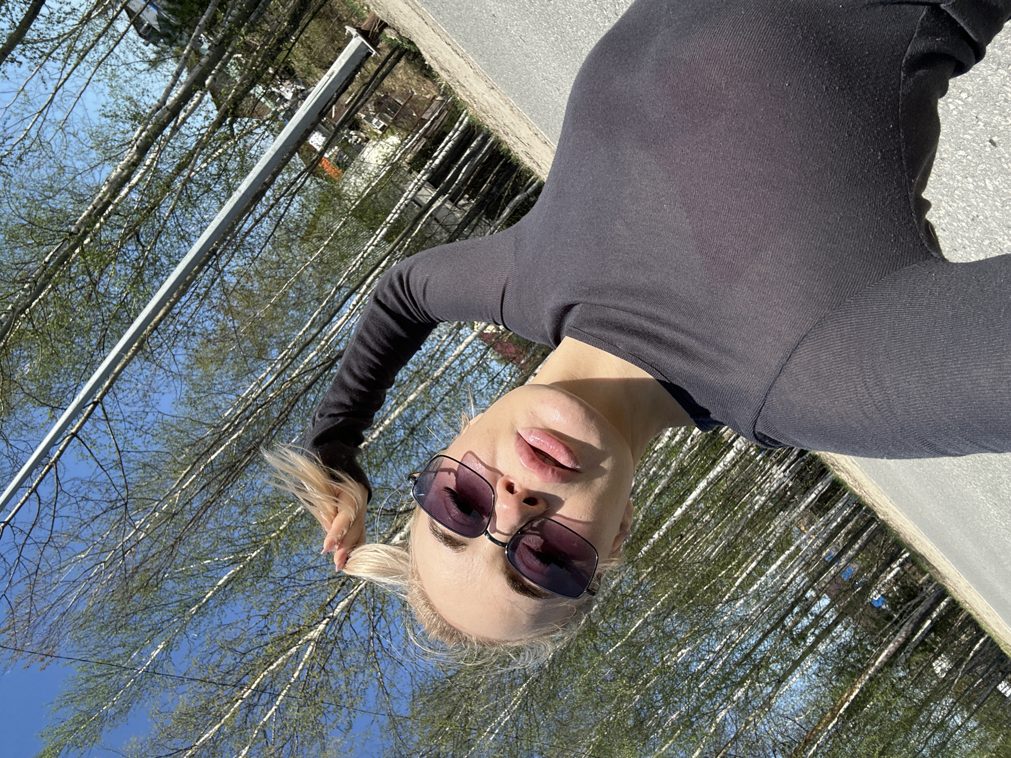 A woman with fair skin and light blonde hair is shown from a low-angle, selfie perspective against a backdrop of birch trees. She is wearing a dark, form-fitting, long-sleeved shirt and dark sunglasses with rectangular frames. Her hair is partially pulled back, with a loose strand held in her hand near the upper left corner of the frame.
In the background, numerous birch trees with light bark and green foliage fill the frame, interspersed with a blue sky. In the upper part of the frame, fragments of buildings are visible among the trees. A metal pole runs diagonally across the upper left quadrant. The ground appears to be paved.
Based on the location specified, Высокий, Russia, the prevalence of birch trees in the background is consistent with the typical landscape of the region.