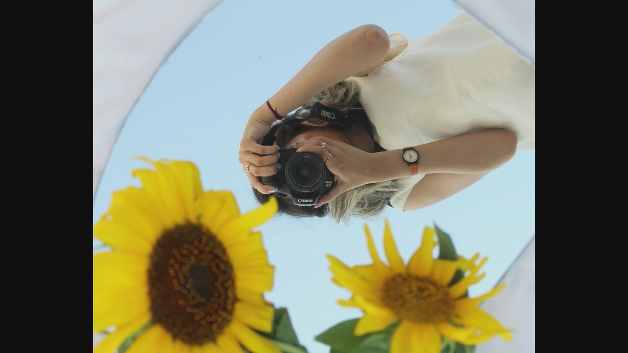 In a reflection in a circular mirror, a person's arms and torso are visible as they hold a black Canon camera to their face, taking a photograph. The person is wearing a cream-colored top, a brown watch with a white face on their left wrist, and a thin red string bracelet on their right wrist. Two bright yellow sunflowers, one larger and out of focus in the upper left, and a smaller one in the lower left, are reflected in the mirror alongside the person. The background of the reflection is a clear blue sky. The scene is not set in a specific city or known location.