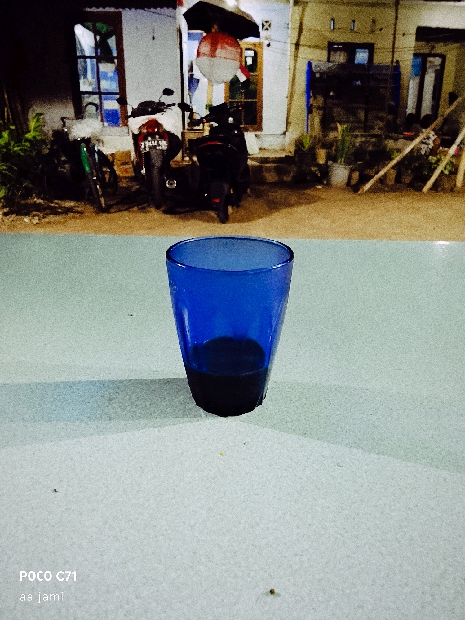 A blue glass is placed on a light-colored textured surface. The glass is partially filled with a dark liquid. In the background, a house with a white facade and wooden elements is visible. Two motorcycles are parked in front of the house. The lighting suggests it is nighttime. The image also contains a watermark that reads "POCO C71 aa jami" in the lower-left corner.