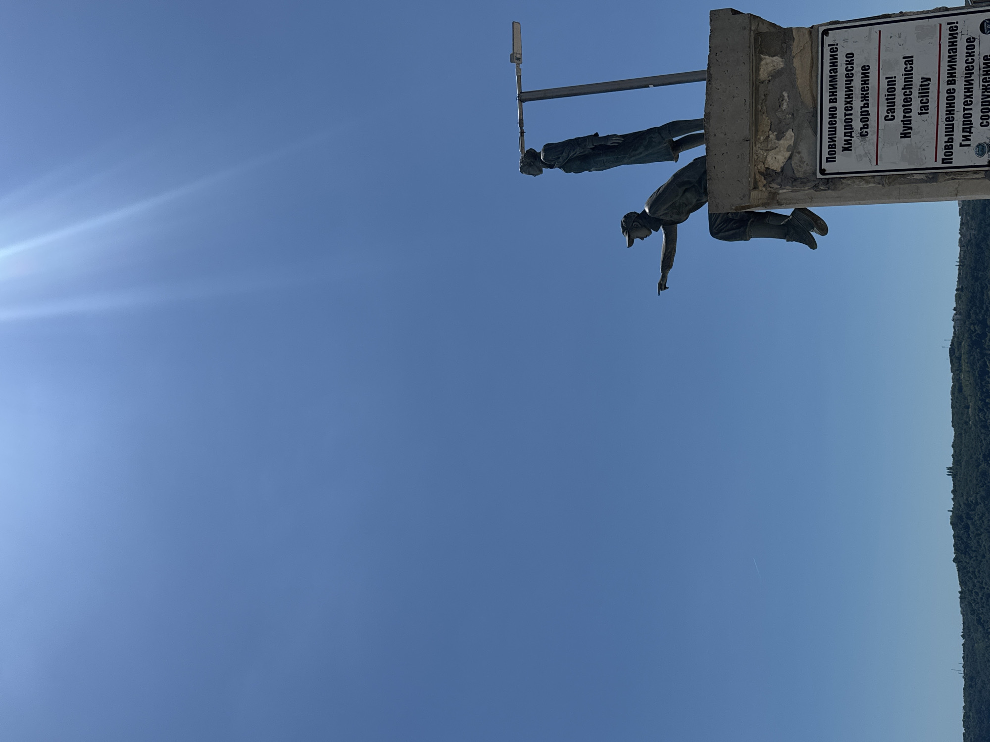 A bronze statue depicts two figures, one seated and pointing to the left, the other standing. They are positioned on a concrete structure against a clear blue sky. A sign is attached to the concrete structure with text in both Cyrillic and English, reading "Повышеное внимание! Гидротехническо съоръжение" and "Caution! Hydrotechnical facility". The background features a distant, tree-covered hillside. The sun is visible as a bright light source at the top of the image, casting lens flares.
