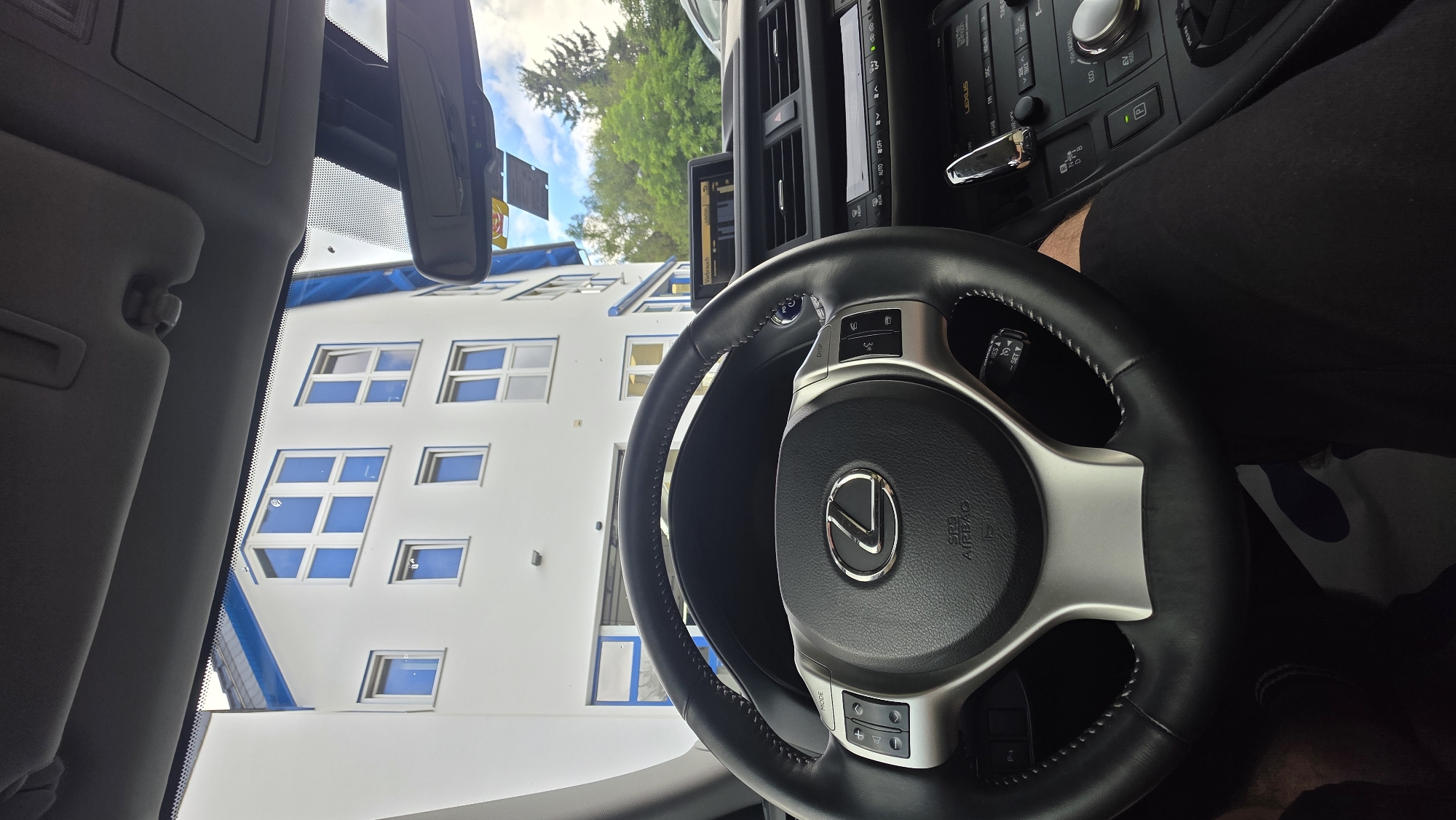 The image is taken from inside a Lexus vehicle, focusing on the steering wheel and dashboard. A building with white walls and blue trim is visible through the windshield, with multiple windows. The sky is cloudy. The steering wheel is black leather with silver accents and the Lexus logo. Controls for the infotainment system and climate control are visible on the dashboard. The driver's legs, covered in black pants, are visible in the lower part of the frame. The scene appears to be set in an urban or suburban environment.