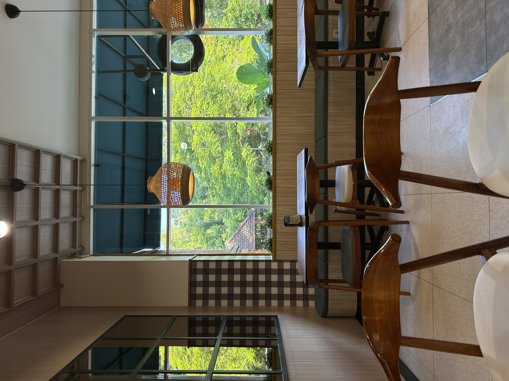 The image shows the interior of a café or restaurant in Kuta, Indonesia. Sunlight streams through large windows, illuminating wooden chairs and tables. The view outside features lush greenery and a glimpse of rooftops. Interior décor includes woven light fixtures and checkered wall accents. No people are visible in the frame.