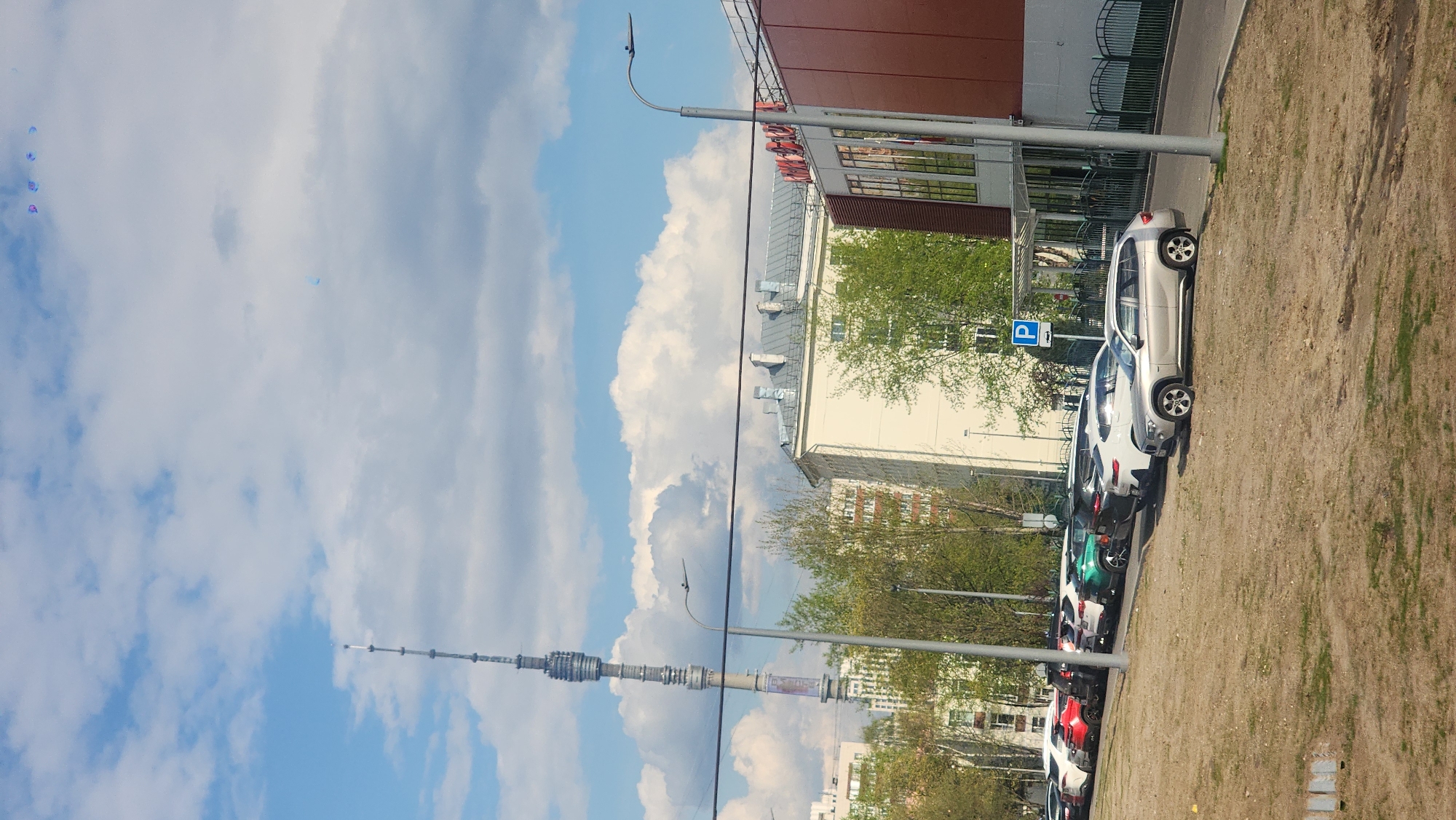 The image presents a scene in Unknown, Unknown featuring a clear blue sky partially covered with fluffy white clouds, with the Ostankino Tower visible in the distance. In the foreground, a row of parked cars of various colors, including silver, white, green, and red, lines the edge of a patch of dirt. A lamppost stands near the parked cars, and a building with red and white facades and grey roofing is also visible. A parking sign with a blue 'P' marks a parking area. Trees with green leaves suggest a springtime setting. Wires traverse the sky.