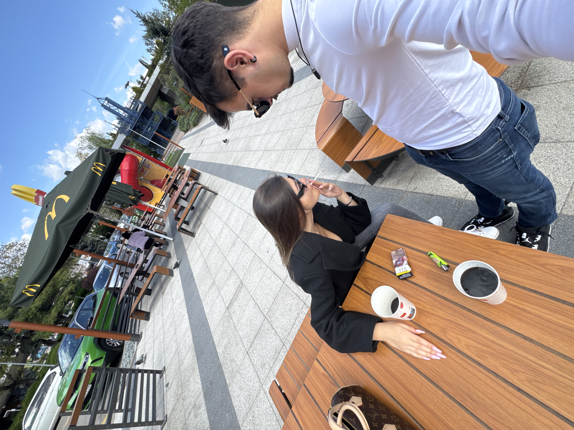The image captures an outdoor scene likely at a McDonald's in Sofia, Bulgaria, as indicated by the McDonald's infrastructure visible in the background. A couple occupies the frame. A man, positioned toward the upper right, wears a white long-sleeved shirt and jeans, with sunglasses partially perched on his head. A woman sits at the table, wearing a black blazer. She is smoking a cigarette. On the wooden table, two coffee cups, a lighter, and a pack of cigarettes are visible, along with a Louis Vuitton handbag. A green car is parked in the background. The setting suggests an urban, public dining area, with visible street-level infrastructure and clear sky above.