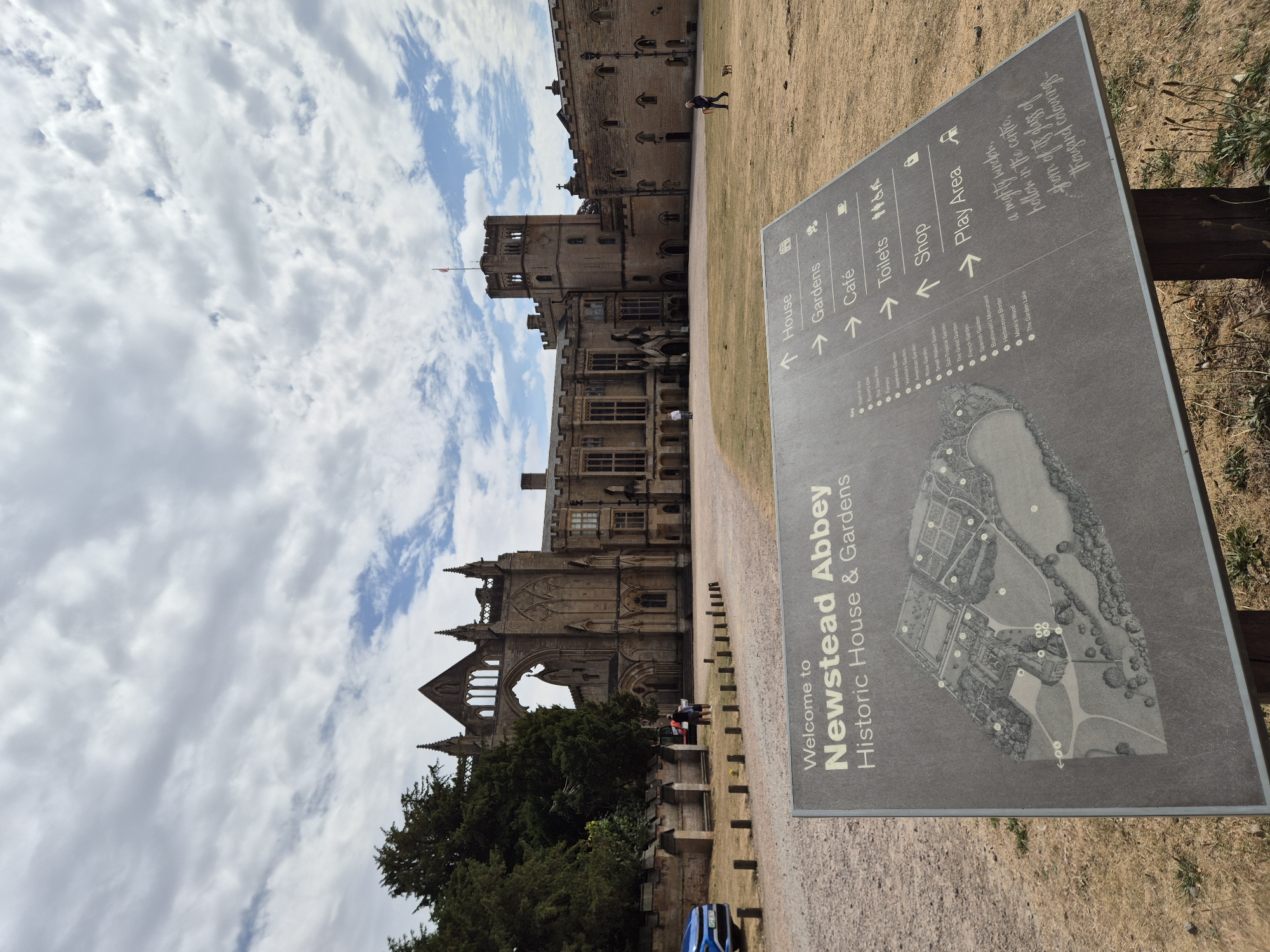 The image depicts Newstead Abbey, a historic house and gardens located in the borough of Gedling, United Kingdom. In the foreground, a dark gray directional sign displays a map of the grounds and lists various amenities such as the House, Gardens, Cafe, Toilets, Shop, and Play Area. The sign also includes a key to specific garden features and a quote. Behind the sign, a gravel path leads towards the abbey buildings. The abbey itself is a large, stone structure with a prominent tower and Gothic architectural elements, parts of which appear to be in ruins. The grounds are covered in dry, yellow grass, indicating a warm season. In the distance, a few individuals are visible walking on the grounds, and a dog is being walked by a person. A blue car is parked on the left side of the image. The sky above is cloudy with patches of blue.