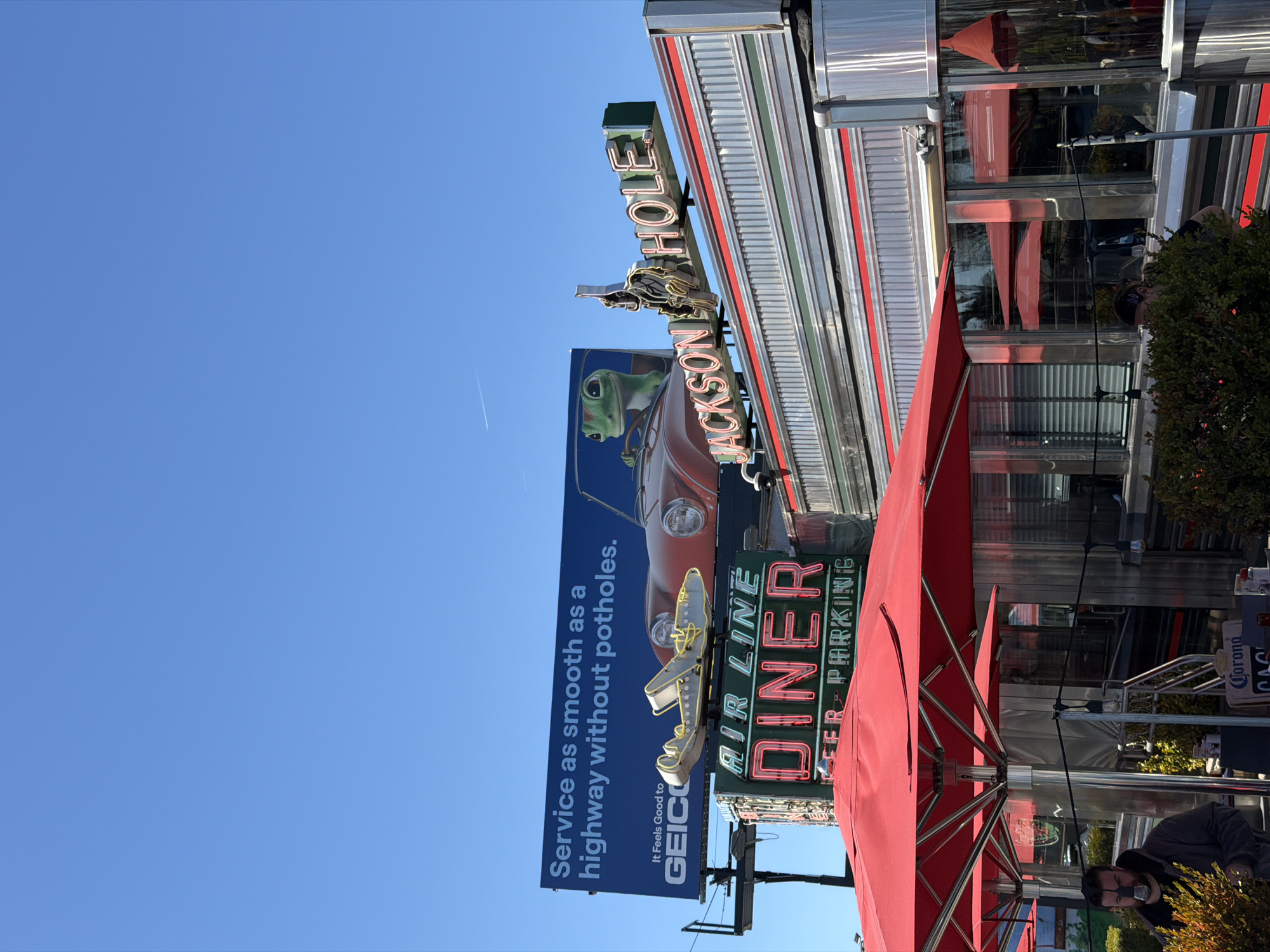 The image shows a street scene with a clear blue sky in New York City. A prominent feature is a billboard advertising GEICO insurance with the slogan, "Service as smooth as a highway without potholes." The billboard is partially obscured by signs for the "Airline Diner" and "Jackson Hole" restaurant. The diner's exterior is visible, featuring a retro design with red and silver accents. A red umbrella partially covers the diner's entrance. A man is visible in the lower-right corner of the image. The general atmosphere suggests a sunny day in a busy urban area.
