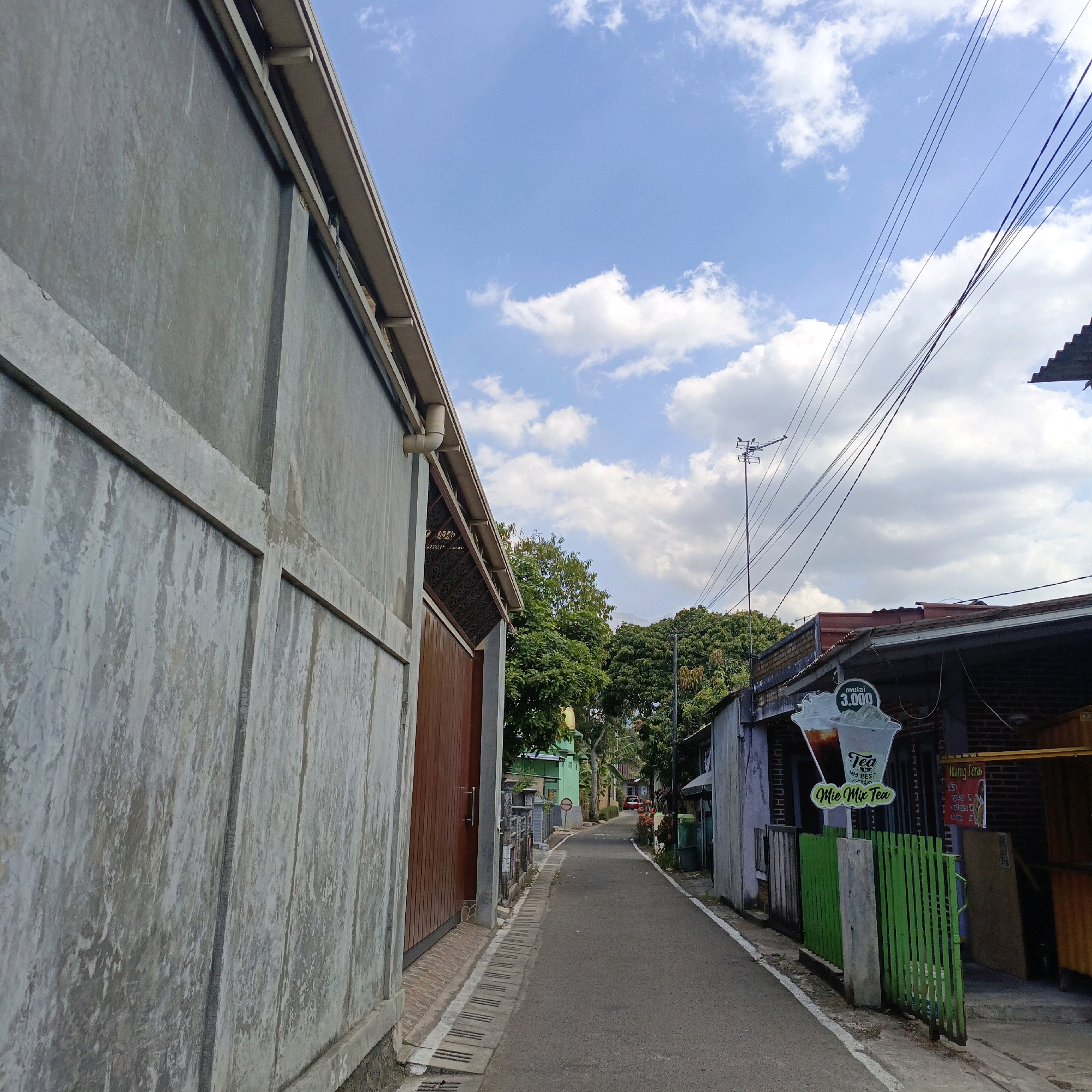 The image captures a narrow street in Salatiga, Indonesia. On the left, a large, unfinished concrete wall dominates the foreground, featuring visible horizontal and vertical concrete beams and a textured, weathered surface. To the right of this wall, a brown wooden door is visible, leading into a covered area with intricate metalwork.
The street itself is paved with asphalt and is bordered by a drainage channel with concrete grates on the left. On the right side of the street, a row of buildings lines the path. These buildings have various facades, including some with grey walls and others with darker brickwork. A green wooden fence stands in front of one of these structures, and a sign advertising "Tea My Best" and "Mie Mie Tea" with a price of "mulai 3.000" is prominently displayed. Further down the street, trees and more buildings are visible, with a red car parked in the distance. Overhead, power lines crisscross the bright blue sky, which is dotted with white, puffy clouds. A television antenna is also visible on a pole. The overall impression is of a residential or commercial street in a Southeast Asian urban environment.