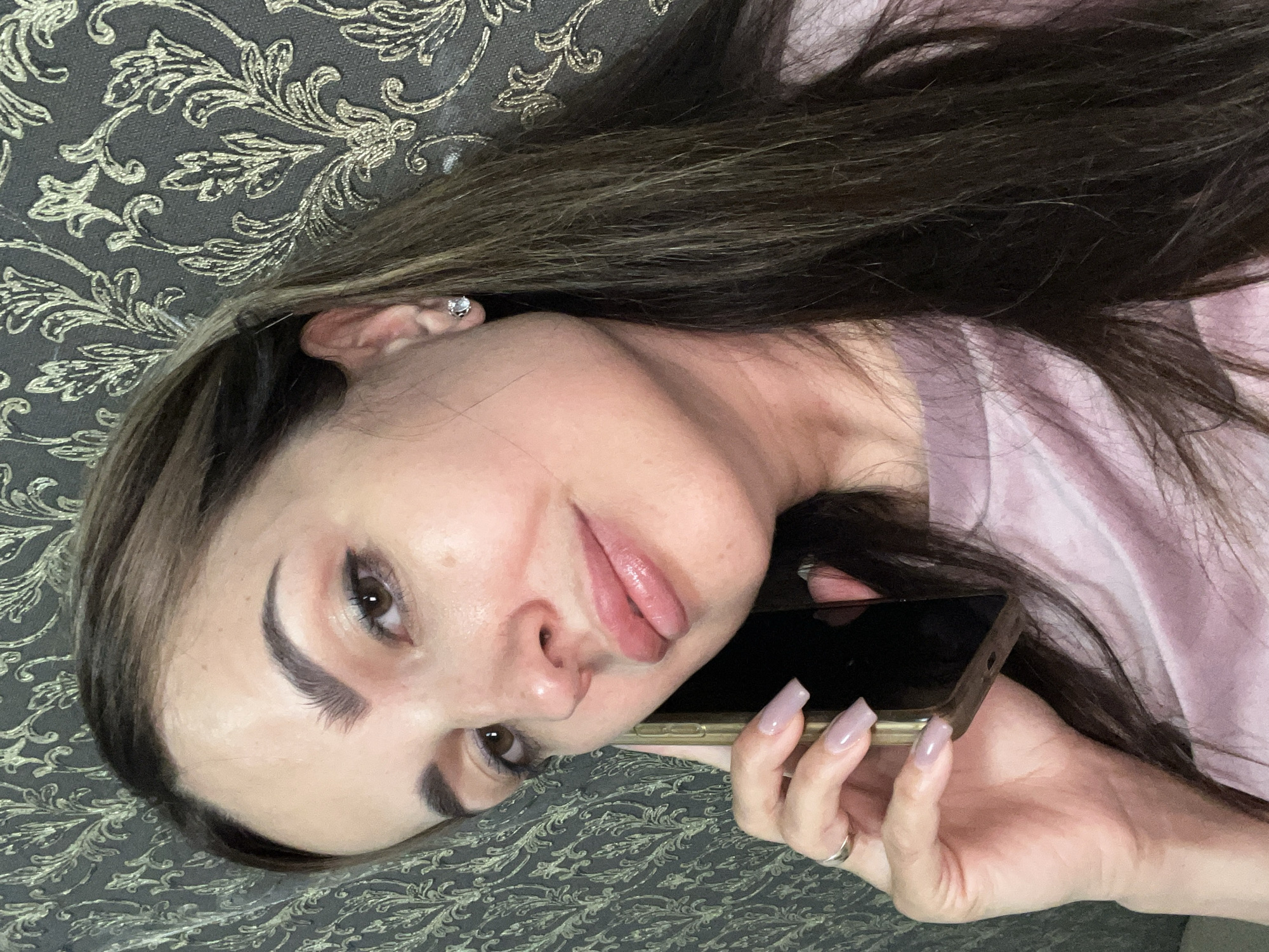 A medium shot captures a woman holding a gold smartphone to her left ear while looking directly at the camera. She has long, dark brown hair with lighter streaks, a light pink shirt, and delicate makeup. Her nails are painted a pale lavender, and she wears a silver ring on her left hand and a small stud earring in her visible left ear. The background features a dark, textured wallpaper with a metallic gold floral pattern. The scene appears to be indoors, with no specific city or location details evident.