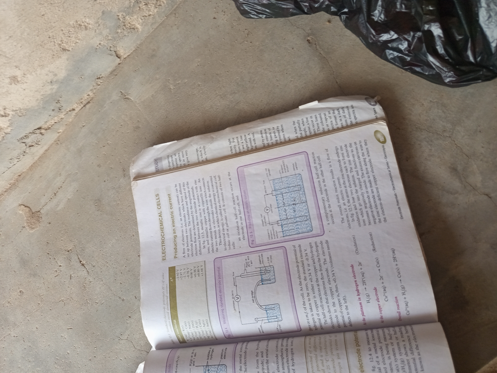 The image displays a page from a textbook open on a textured, concrete surface. The textbook page contains text and diagrams related to electrochemistry. The concrete surface appears to be an outdoor environment, possibly a walkway or ground, with some dust and debris. The lighting suggests it's daytime. There are no people or animals visible in the image. The focus is on the book and its contents. The surrounding environment is out of focus, but the rough texture of the concrete is discernible. The book is open to a section titled "ELECTROCHEMICAL CELLS" and details topics like "Producing an electric current," "Measuring standard electrode potential," and includes diagrams of electrochemical cells. One diagram shows a setup with a copper electrode and a platinum electrode connected by a salt bridge and a voltmeter. Another diagram illustrates a cell with a copper rod and a zinc rod. The text discusses electrode potentials, half-cell reactions, and electron flow. The page number 137 is visible.