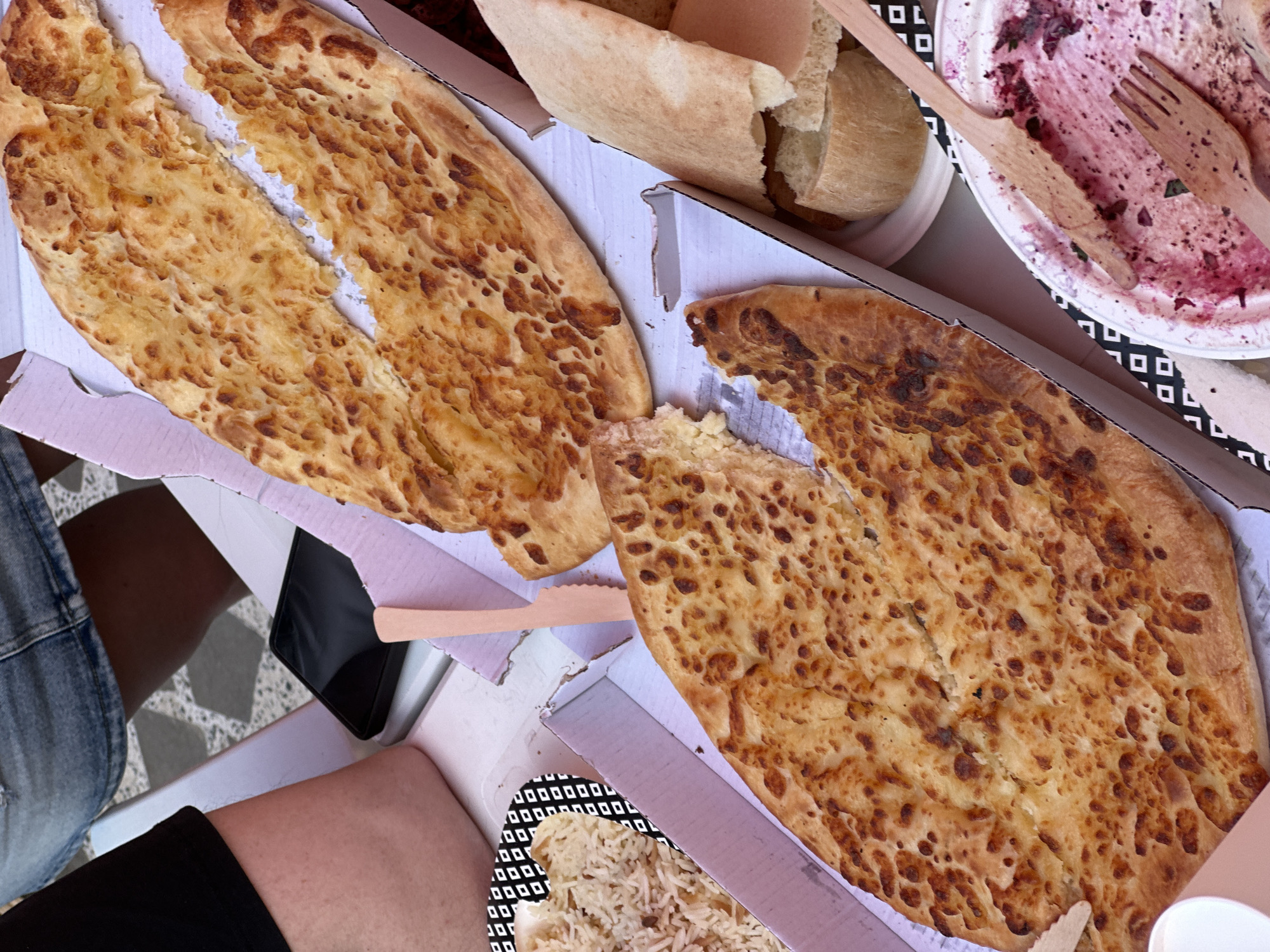 An overhead view captures an informal meal, likely taking place outdoors or in a casual setting. Two prominent, golden-brown pieces of Georgian cheese bread (khachapuri) are displayed in cardboard takeaway boxes; one is split open down the middle, revealing a cheesy interior, while the other shows a bite taken out of it.
Various other food items and accessories contribute to the scene: pieces of plain bread are visible in another container, and a plate of rice (possibly pilaf) with a distinctive black and white geometric pattern around its edge is also present. The casual nature of the meal is highlighted by the use of disposable items, including an empty white paper plate heavily stained with purple berry or fruit residue, and two wooden utensils (a fork and a spoon/knife) resting on it.
Parts of two individuals are visible, including a leg in denim shorts or jeans and another leg with bare skin next to a dark fabric. A black smartphone is placed on a surface next to a white paper cup. The ground or floor beneath the eating area features a grey and white geometric tile pattern. The bright lighting indicates it is daytime. The overall impression is a relaxed, communal dining experience, possibly a picnic or shared meal in Tbilisi, Georgia, as suggested by the traditional khachapuri. No legible text is visible in the image.