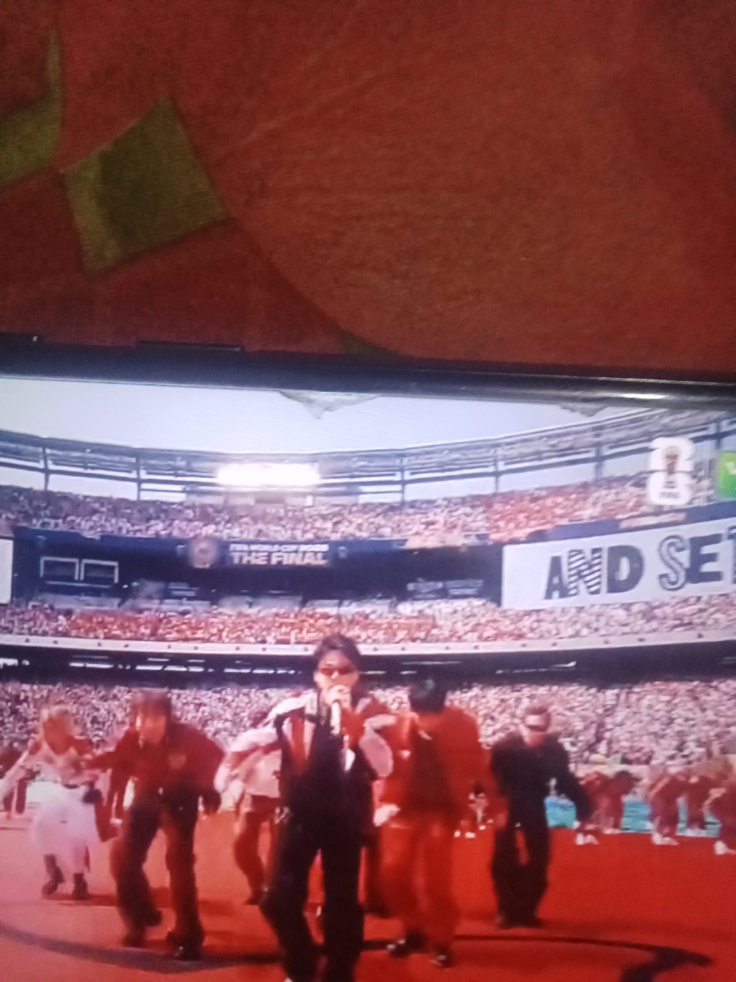 The image depicts a screen displaying a live event, possibly a concert or sporting event, held in a large stadium. In the foreground, several individuals are visible on a red stage. One person in the center is holding a microphone and appears to be performing. Other individuals are dressed in various outfits and are positioned around the performer.
In the background, a stadium filled with spectators is visible. A large banner with text is displayed across the stadium, part of which reads "THE FINAL". Another banner displays "AND SE". The overall scene suggests a large-scale public event taking place within a stadium environment.
The image does not contain any information that would allow for its location to be identified as Buketglumpang, Indonesia.