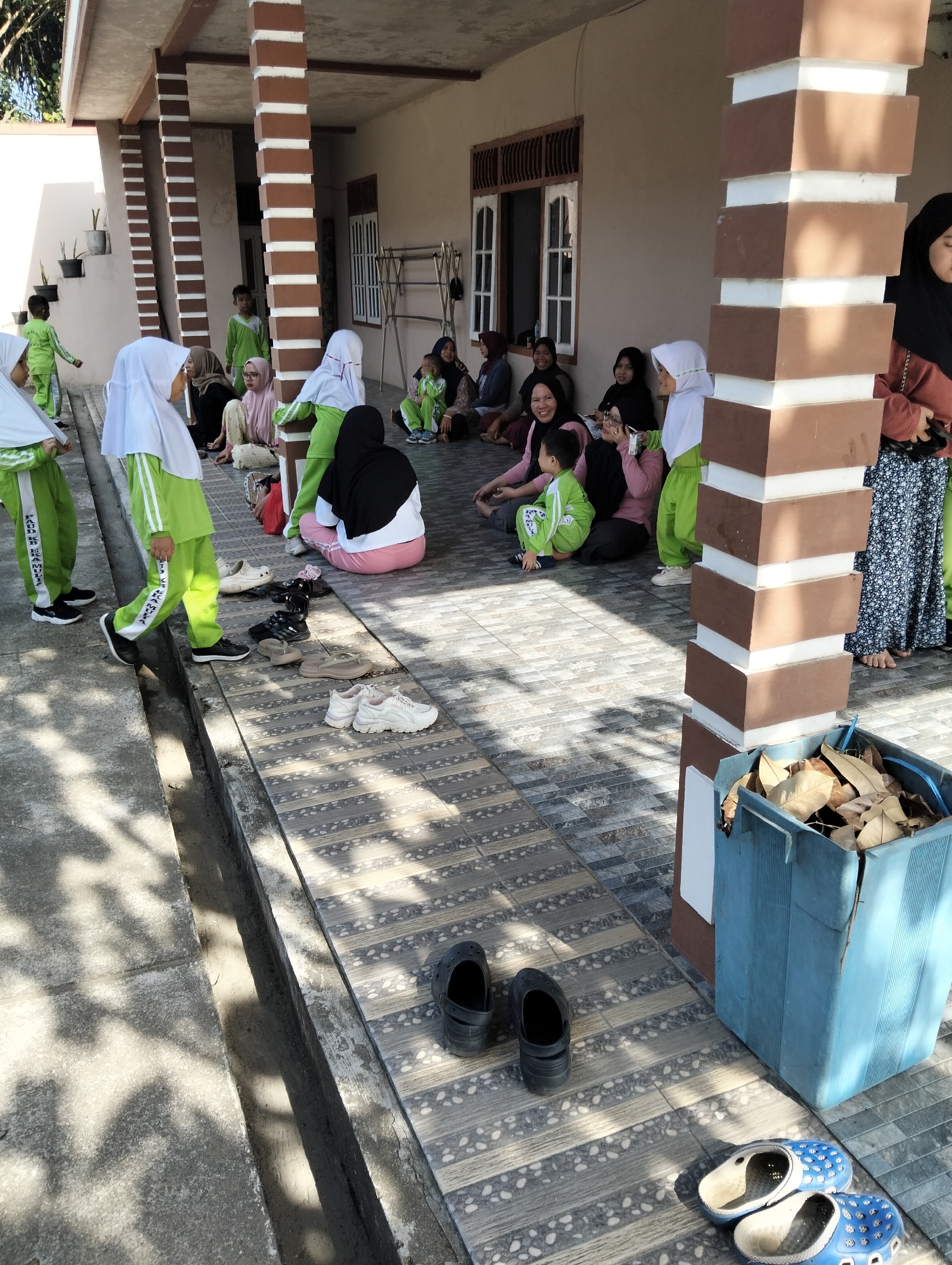 The image depicts a scene in front of a building with a covered patio in Janju, Indonesia. Several people, including adults and children, are gathered on the patio and the tiled floor. Many of them are wearing green uniforms, suggesting they might be students or part of a group. Some individuals are seated on the ground, while others are standing or walking. The environment appears to be outdoors, with sunlight casting shadows on the ground. Several pairs of footwear are visible on the tiled area, including black clogs and white sneakers, indicating that people may have removed their shoes before entering the covered area. A blue plastic bin filled with dry leaves is also present. The architecture of the building features prominent brown and white striped pillars supporting the roof.