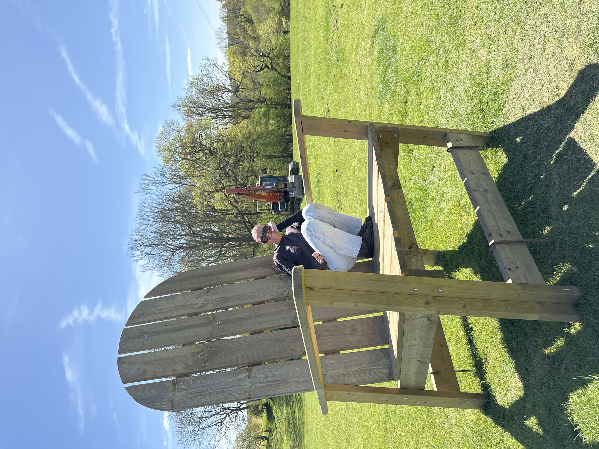 The image captures a sunny outdoor scene in Sevenoaks, United Kingdom, where a person is seated in an oversized wooden Adirondack chair placed on a grassy field. The individual, wearing sunglasses, a dark top, and light-colored jeans, is reclined in the chair, facing the camera. The chair itself is made of light-colored wood and appears to be a novelty piece due to its large size. In the background, there's a green field, trees, and construction equipment, possibly a digger, partially obscured by the trees. The sky is blue with some wispy clouds. A shadow cast by the chair is visible on the grass.