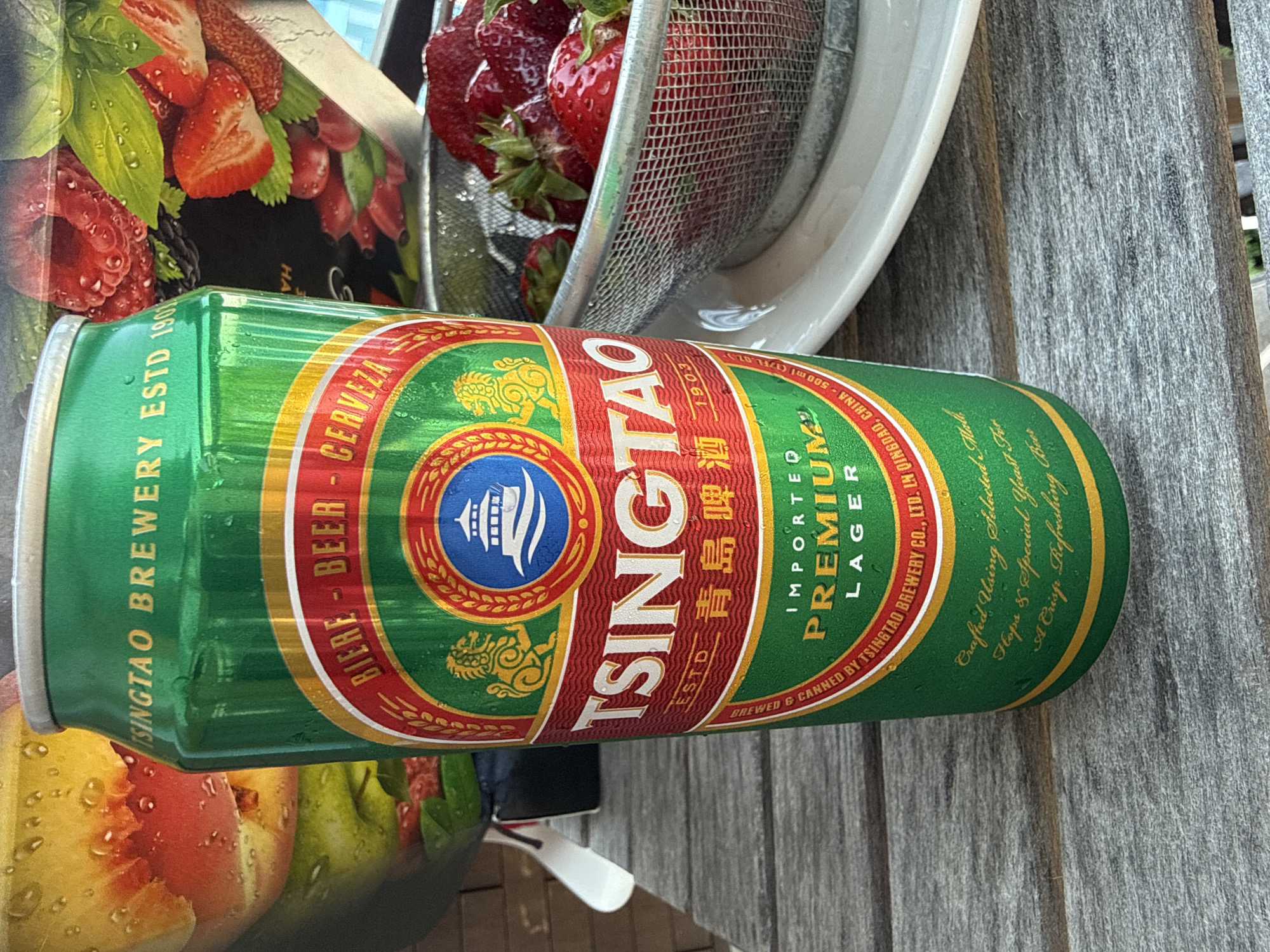 The image shows a green Tsingtao beer can sitting on a weathered wooden table. The can has a prominent label with the brand name "TSINGTAO" in large white letters, along with other text indicating it is a premium lager, brewed and canned by Tsingtao Brewery Co., Ltd. in Qingdao, China. The can is slightly damp, suggesting condensation. To the right of the can, a silver metal colander is filled with fresh strawberries. In the background, out of focus, there is an image of fruits, including apples and berries, on what appears to be packaging or a poster. The scene is outdoors, with the wooden table and blurred background suggesting a casual setting. There is no indication of the city of Sochi, Russia.