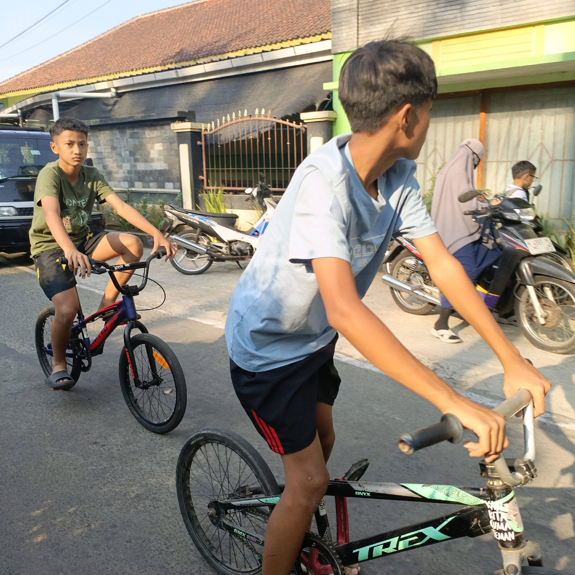 The image depicts two teenage boys riding bicycles on a paved road in Salatiga, Indonesia. The boy in the foreground is wearing a light blue t-shirt and black shorts, while the boy behind him is wearing a green t-shirt and black shorts. They are both looking forward, seemingly engaged in a bicycle ride.
In the background, a woman in a grey hijab and a person on a motorcycle are also present, indicating a public street scene. The surroundings suggest a residential area with houses visible, one with a red tiled roof and another painted green. The lighting indicates it is daytime, likely late afternoon due to the warm, angled sunlight casting shadows. The road surface is asphalt, with some minor wear and tear. The presence of a parked dark-colored vehicle on the left further supports the urban or suburban setting. The overall atmosphere is casual and everyday.