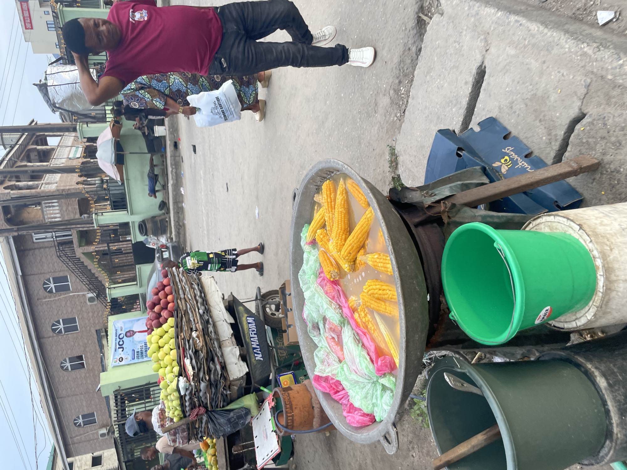 This image captures a vibrant street scene in Ado, Nigeria, depicting everyday life and commerce. The setting is an urban street with multi-story buildings in the background, a concrete sidewalk, and various street vendors. The overall impression is that of a bustling marketplace or a lively commercial area.
In the foreground, a man wearing a maroon t-shirt, dark trousers, and white sneakers stands casually, with one hand behind his head, looking towards the camera. Behind him, another individual in a patterned traditional outfit carries a white shopping bag labeled "DESMOND BOUTIQUE." A young person or child in a green and black patterned outfit is seen walking further down the street.
The most prominent objects in the immediate foreground are a large, grey metal basin filled with water, containing numerous ears of yellow corn. Some items wrapped in pink and light green plastic are also floating in the basin. Adjacent to this, there's a bright green plastic bucket with the word "AMBER" on it, and another dark bucket containing a wooden stick and a metal utensil. A blue cardboard box with a bee logo and "ruitways" is visible on the ground nearby.
Further into the scene, a street vendor's stall or cart is laden with an abundant display of fruits, including piles of red apples and green apples (or similar produce). Beside this stall, a motorized tricycle or cart has the name "NAMARYAM" clearly written on its side. An umbrella provides shade in the mid-ground.
The activity primarily revolves around street vending and daily errands. People are walking, and goods are openly displayed for sale. The time of day appears to be daytime, possibly late morning or afternoon, under an overcast sky.
Visible text in the image includes: "DESMOND BOUTIQUE" on a white bag, "NAMARYAM" on a cart, "AMBER" on a green bucket, and "ruitways" with a bee logo on a blue box. A banner in the background reads "READY TO SERVE JCC JOHN CAMPOS OGUNDARE." Other fragmented text like "LOTALGES" is also discernible.